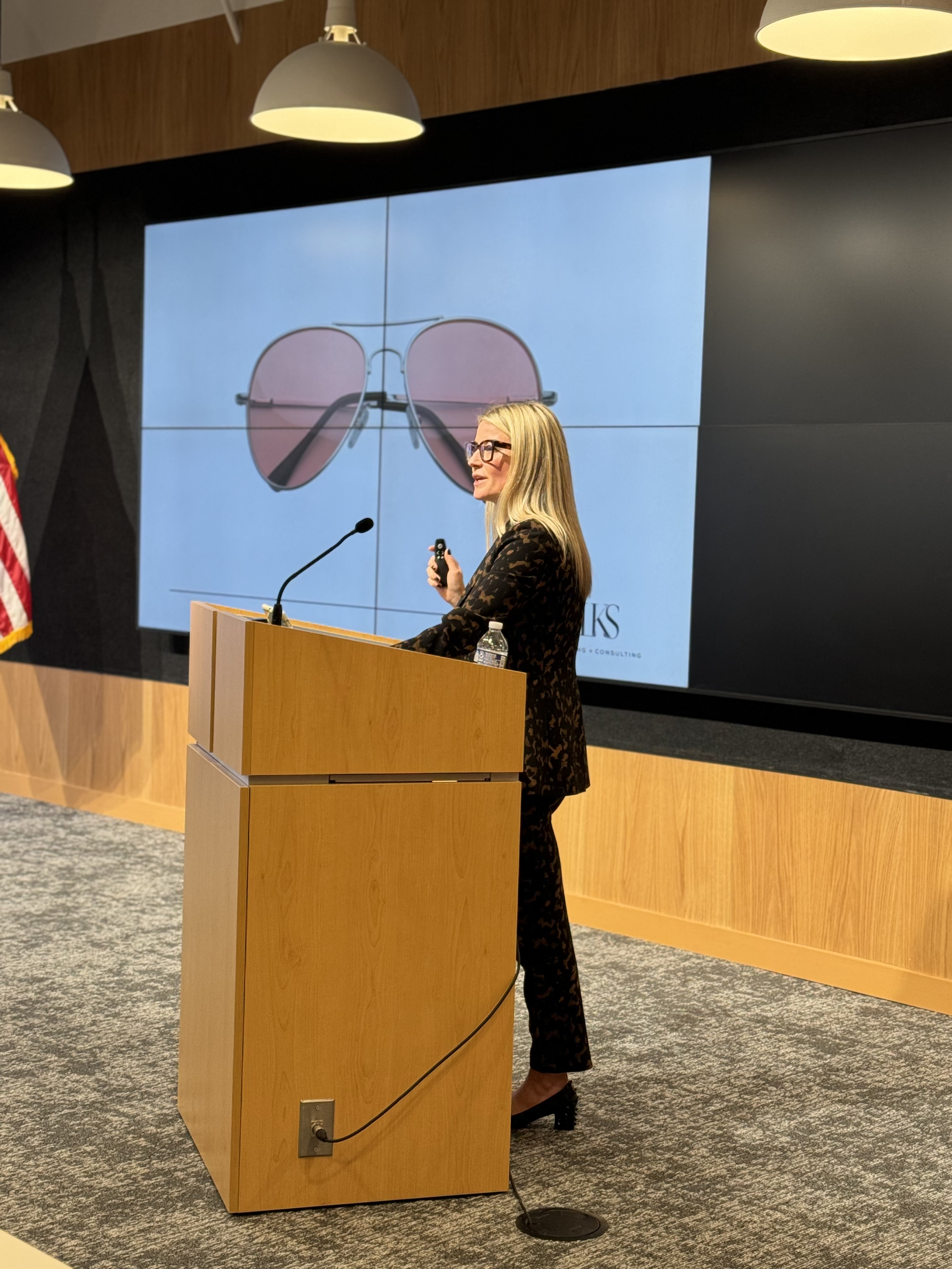 Laura K. Sawyier is giving a presentation at a wooden podium with a bottle of water, microphone, and a remote control. She is speaking in front of a large screen displaying an image of sunglasses.