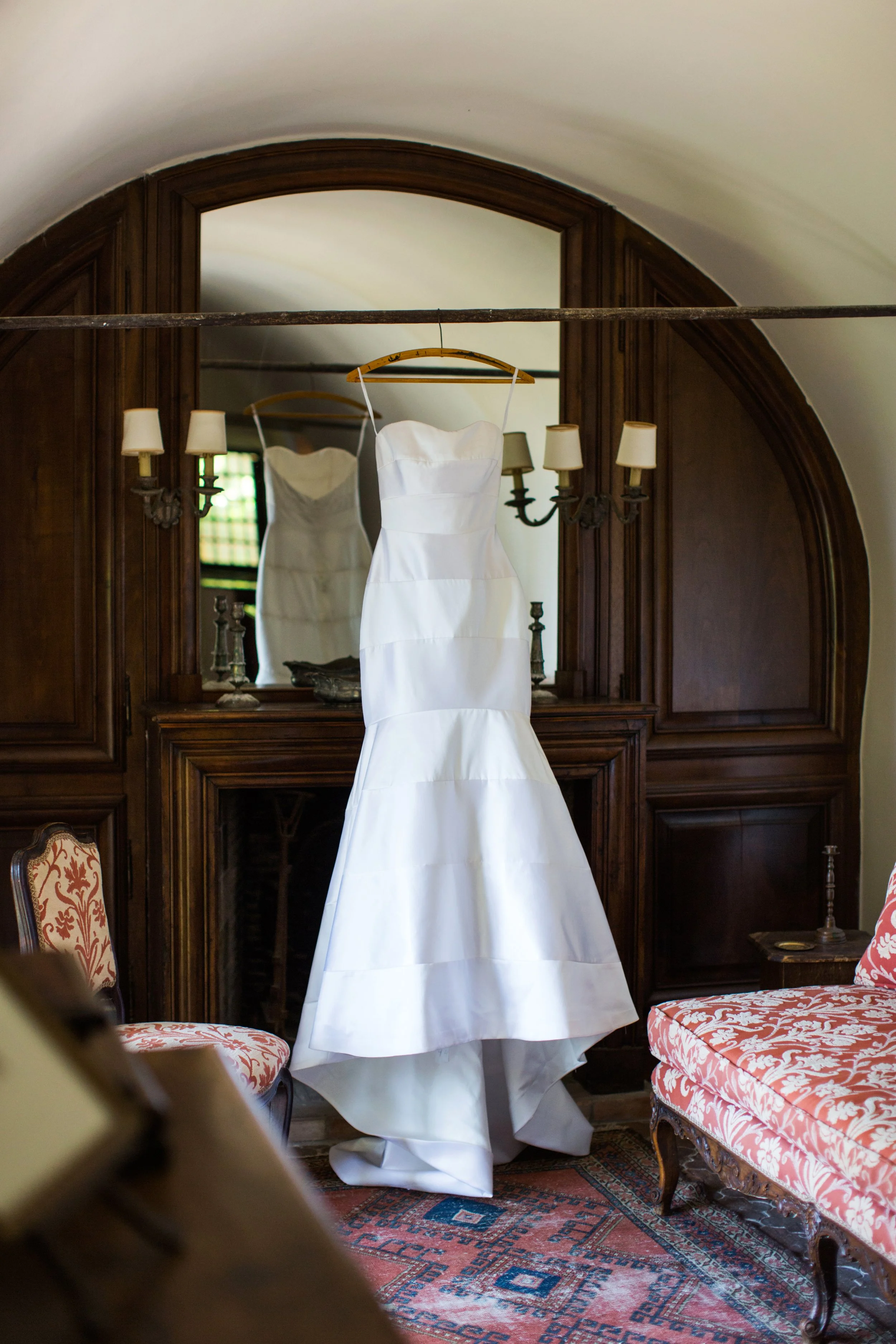 A white wedding dress hanging on a hanger in front of a mirror in a room with dark wooden paneling and furniture.