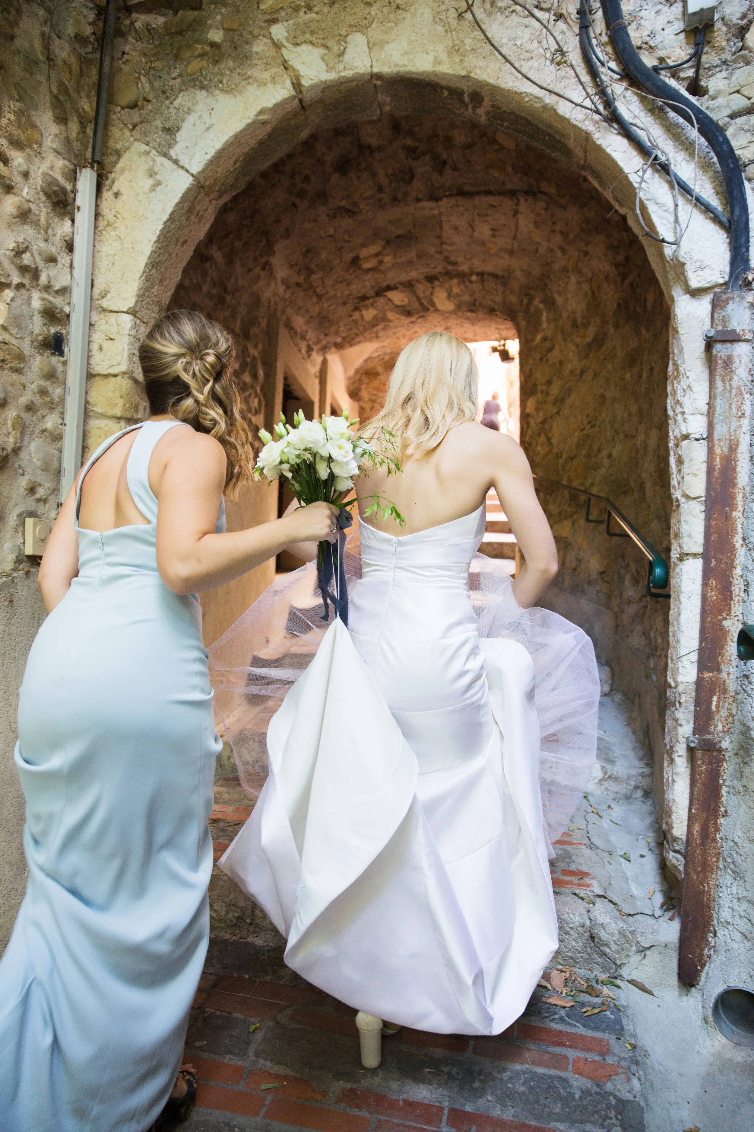 A bride in a white wedding gown walking up a stone staircase, accompanied by a bridesmaid holding a bouquet of white flowers.