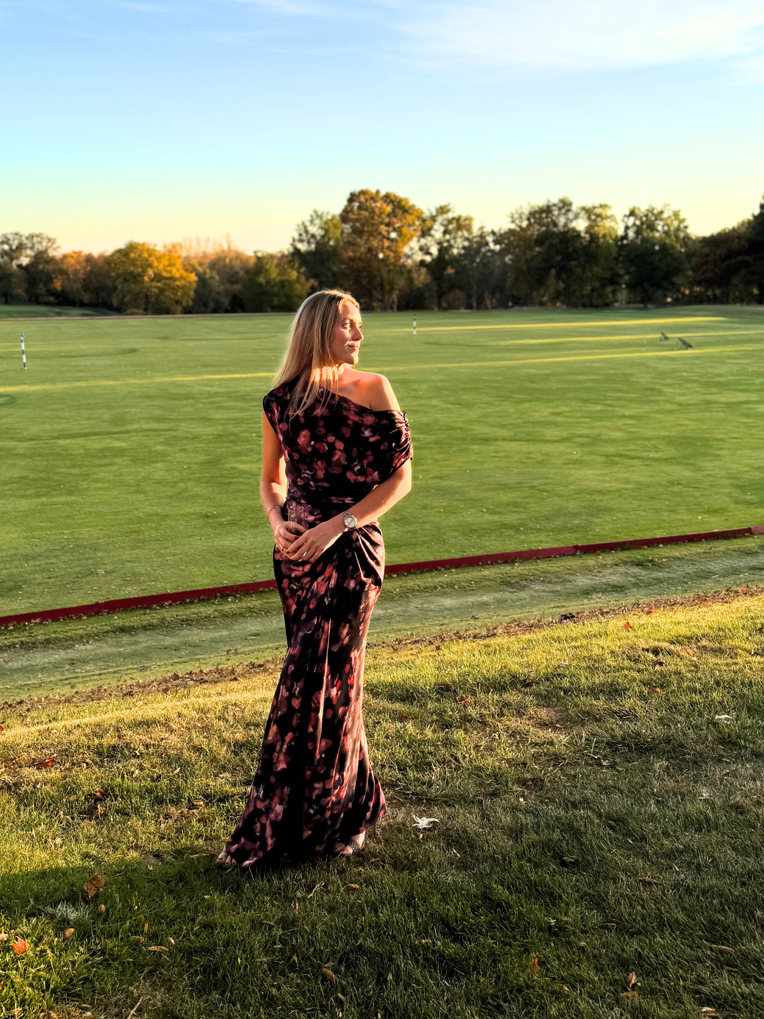 A woman in an off-shoulder floral dress standing on grass near a golf course at sunset.