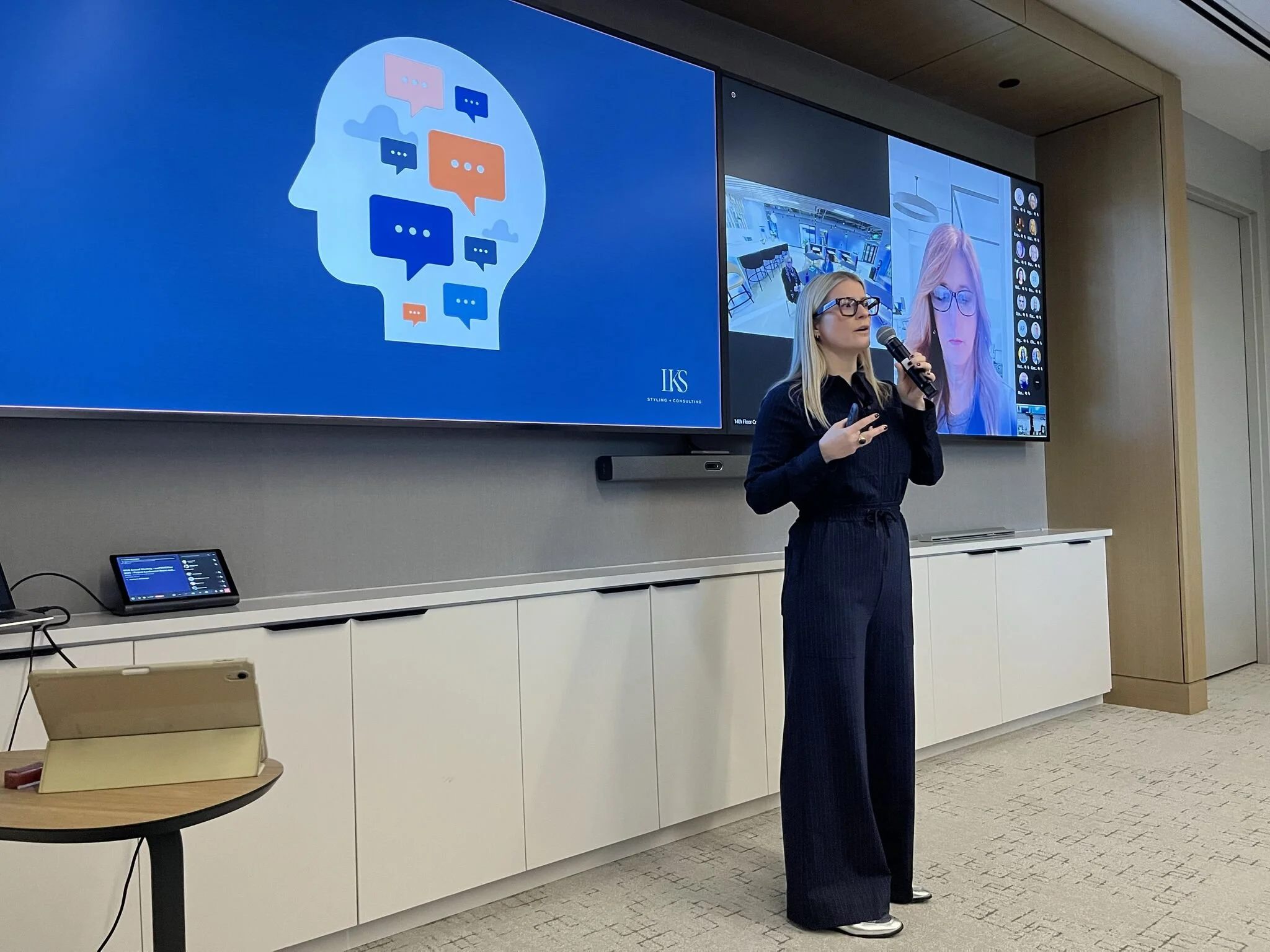Laura K. Sawyier stands in front of a presentation screen, holding a microphone and a remote, giving a talk in a conference room. 