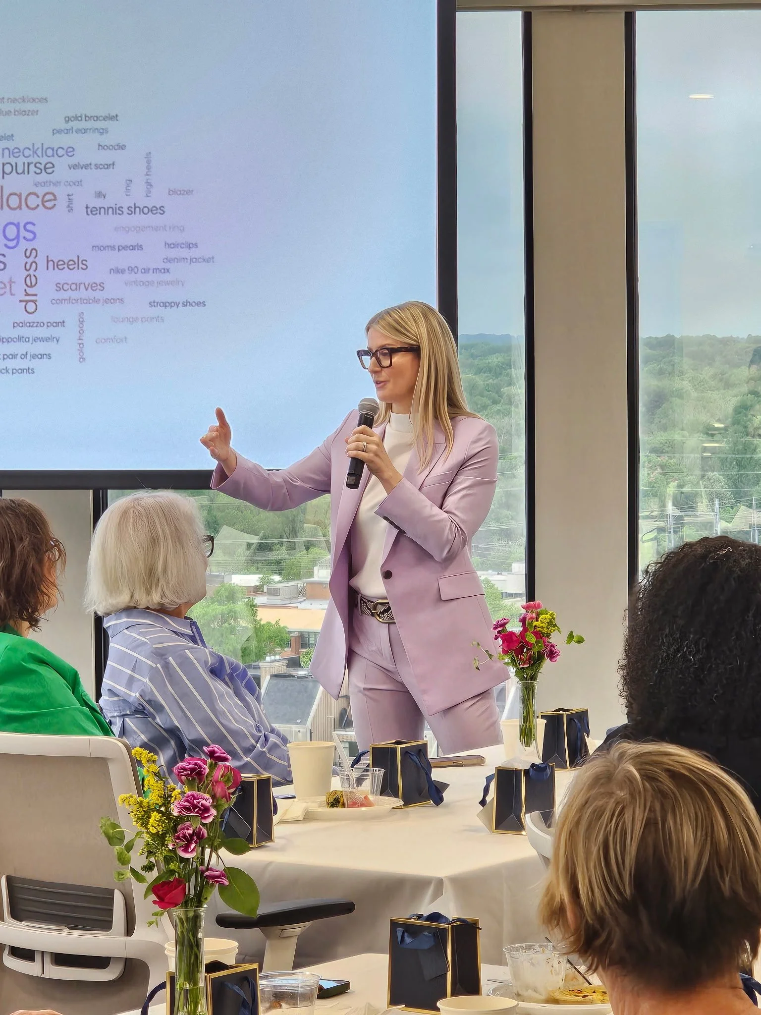 Laura K. Sawyier is giving a presentation to a group of women seated at a round table with flower centerpieces, gift boxes, and food, with a large window showing a green landscape outside.