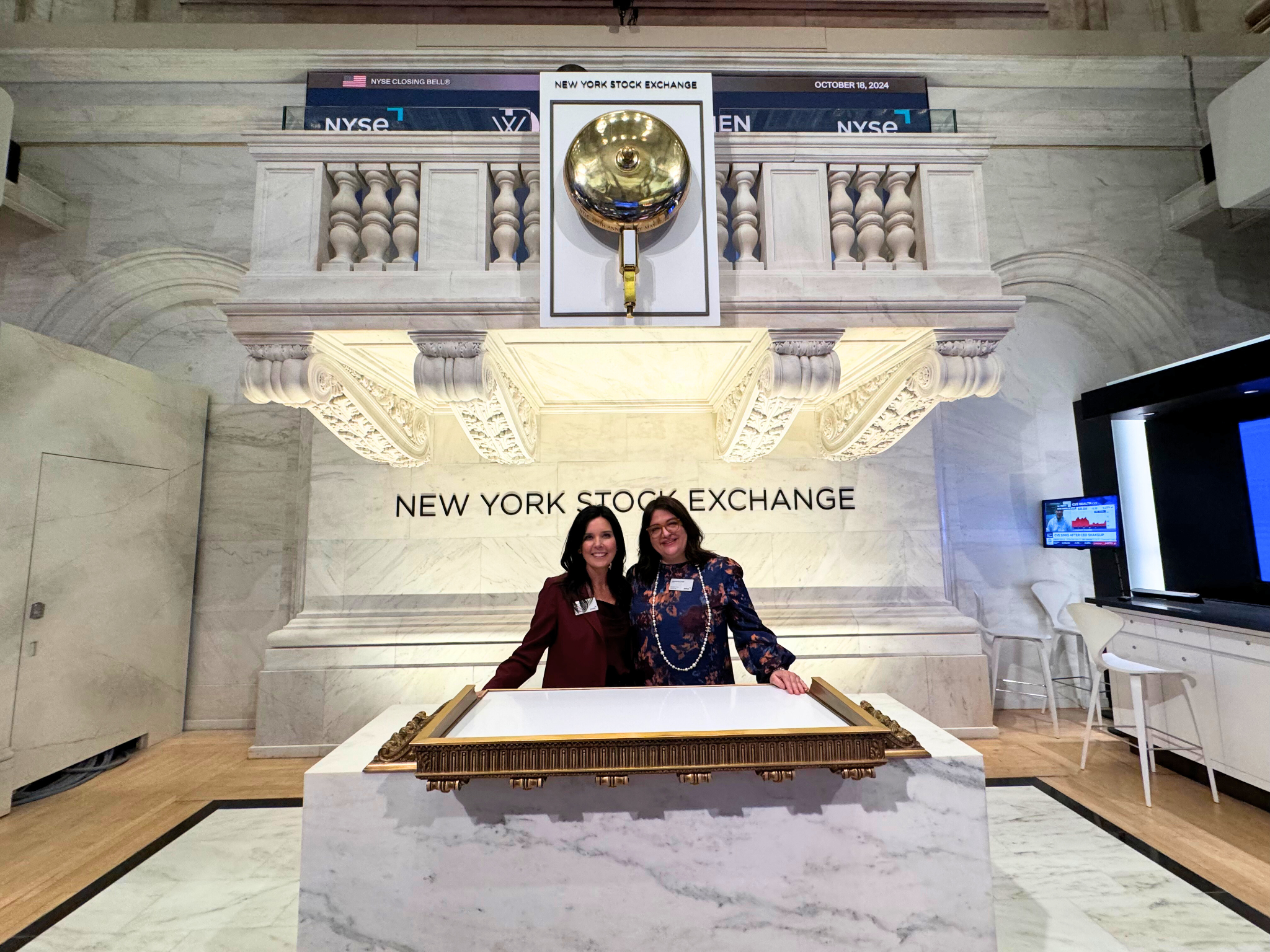 Two women standing behind a marble desk at the New York Stock Exchange, smiling for the photo. The background features a large golden bell and the sign 'NEW YORK STOCK EXCHANGE' on the wall.
