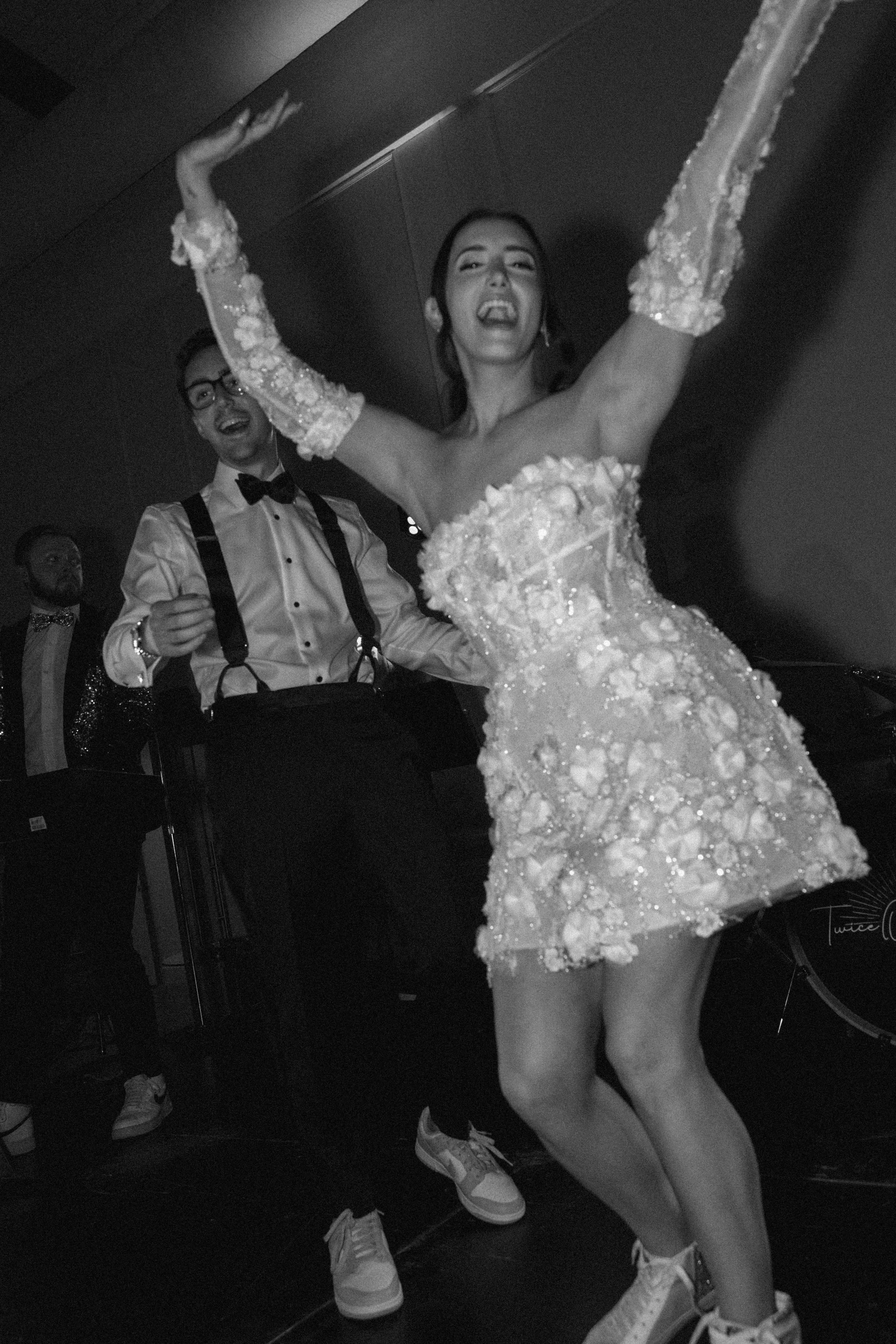 A woman in a short, floral-pattern dress dancing with her arms in the air, smiling, at a celebration event with men in tuxedos in the background.