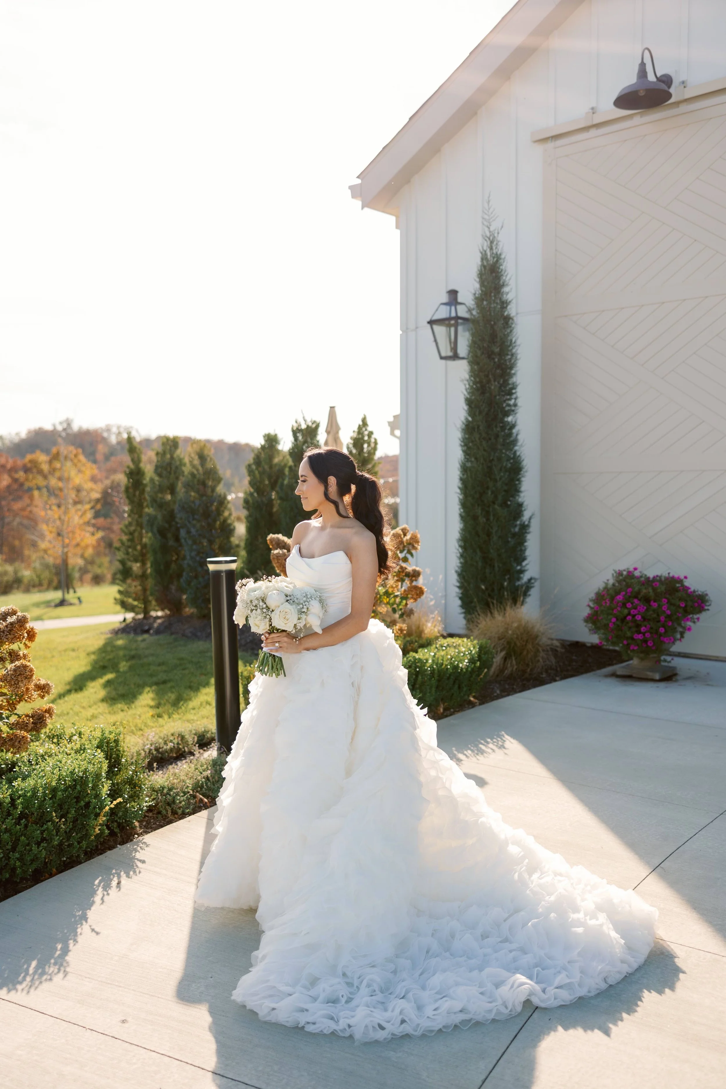 A bride in a white wedding gown holding a bouquet of white flowers standing outdoors near a white building with greenery and trees in the background.