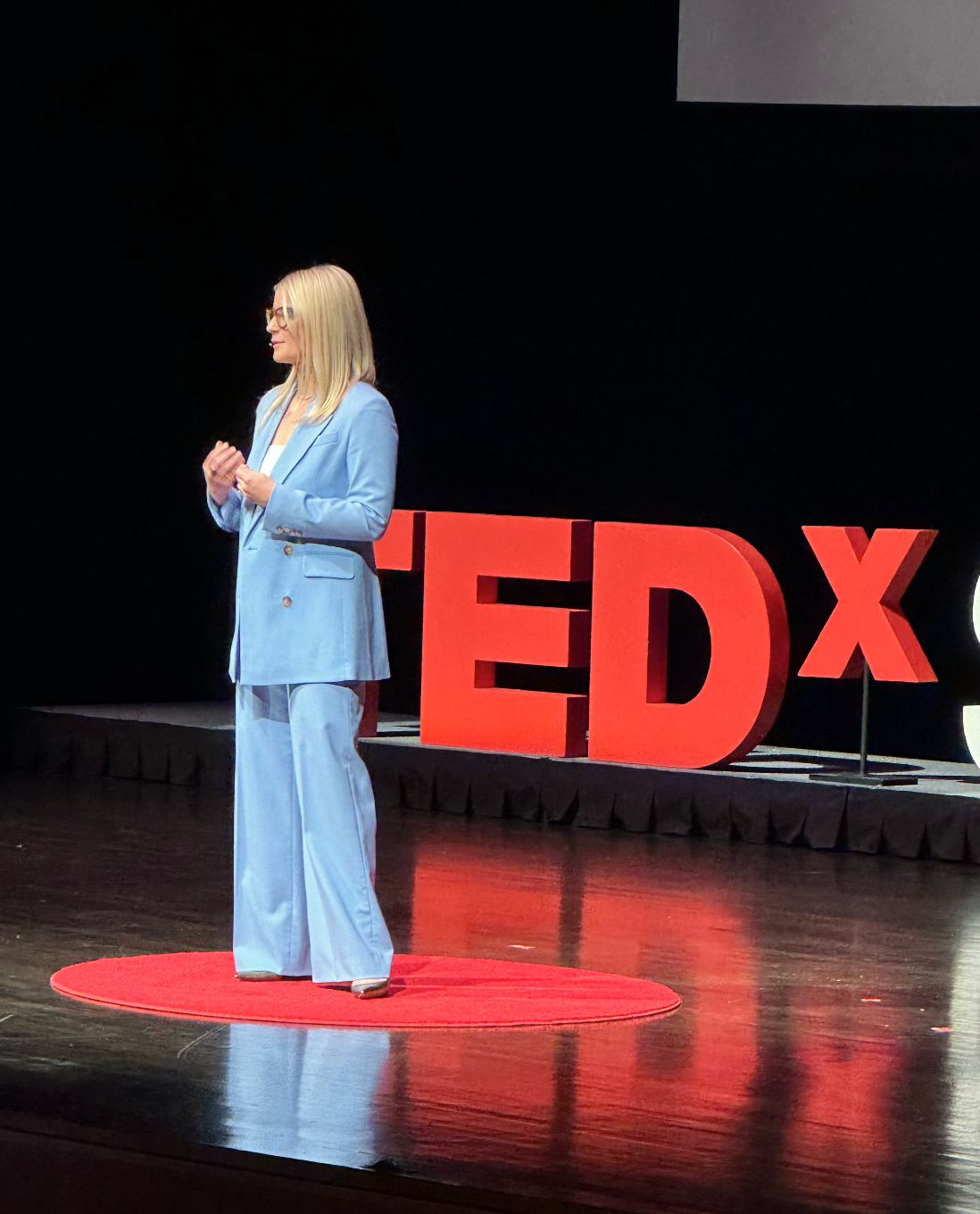 Laura K. Sawyier is speaking on stage at a TEDx event, with large red TEDx letters in the background.