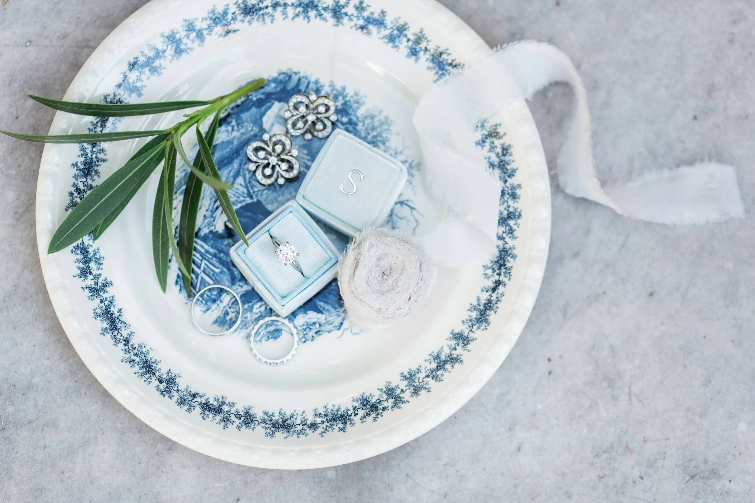 A decorative plate with blue snowflake border holds jewelry including a ring with a large gemstone, a pair of floral earrings, and two wedding bands, with a sprig of greenery and a small ball of white thread on top.