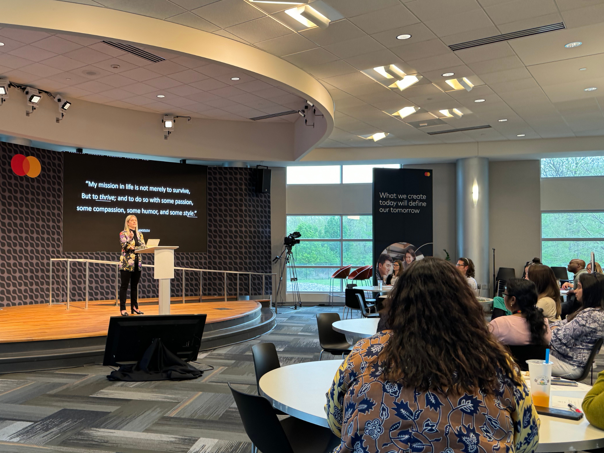 Laura K. Sawyier is giving a presentation on an elevated stage in front of an audience in a conference room. There is a large screen behind her displaying a quote, with windows providing natural light. 
