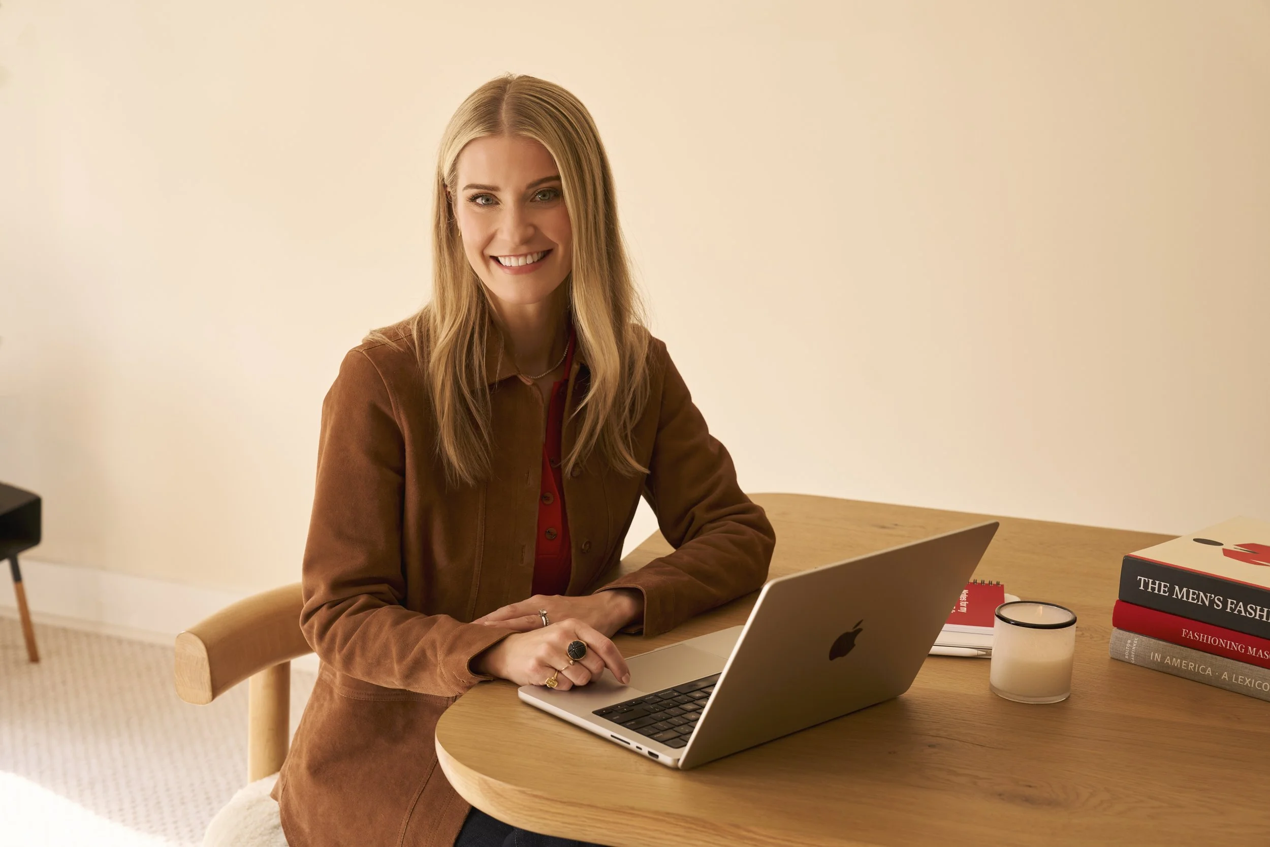 Laura K. Sawyier smiling while sitting at a wooden desk with a silver MacBook, books, a notebook, and a candle in a beige room.