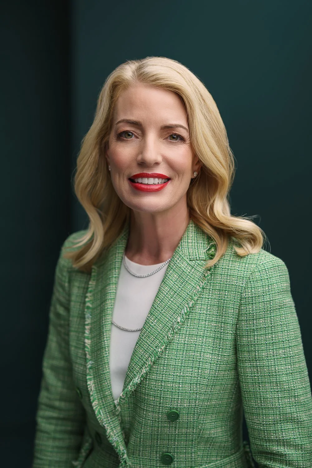 A blonde woman with wavy hair, wearing a green textured blazer over a white top, smiling and standing against a dark green background.