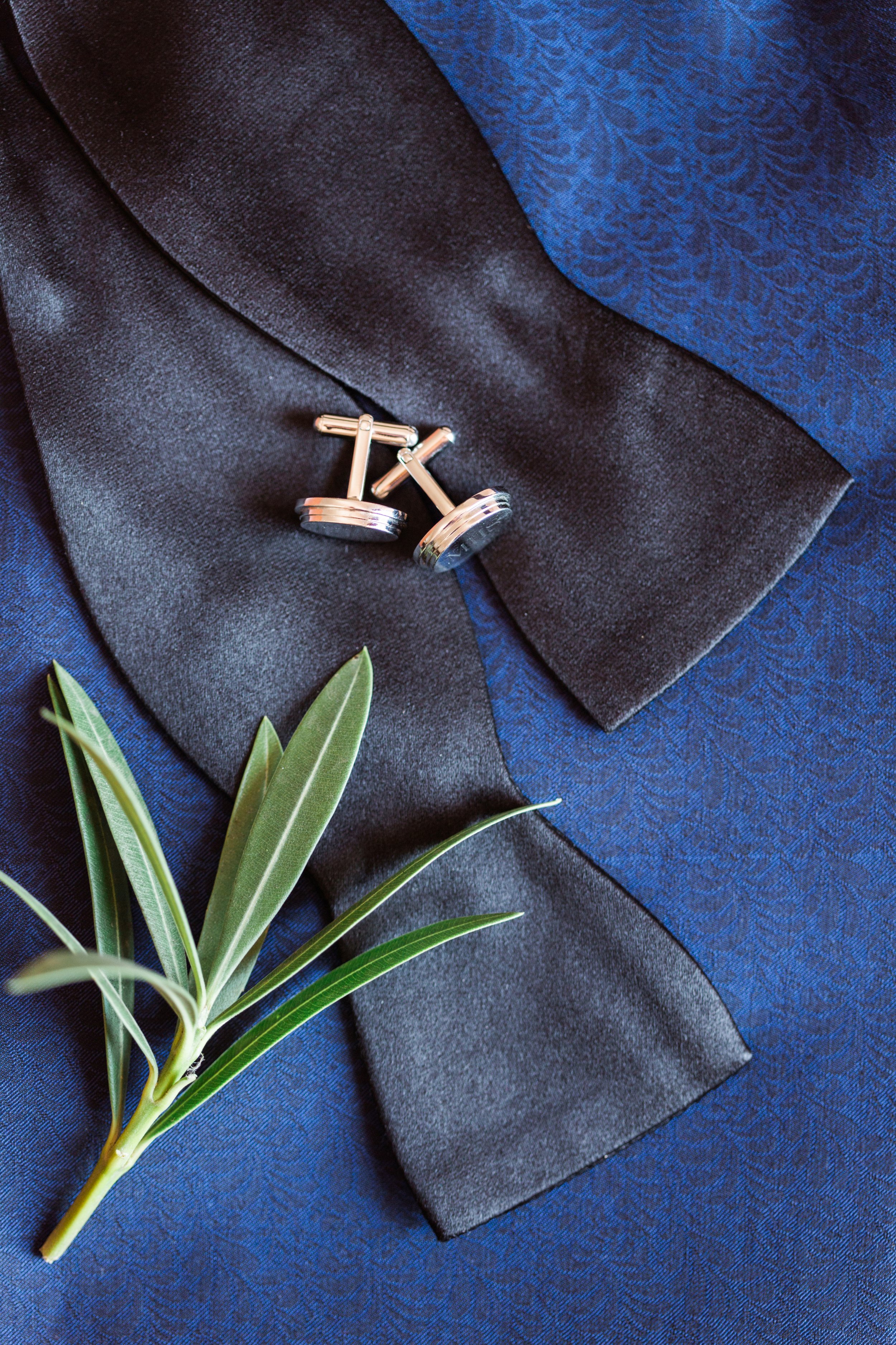 A pair of silver cufflinks placed on a black satin cloth, with a blue textured fabric background and a green leafy branch.