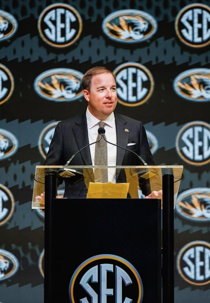 A man in a dark suit and tie standing at a podium with the SEC logo, speaking at an event with multiple SEC logos in the background.