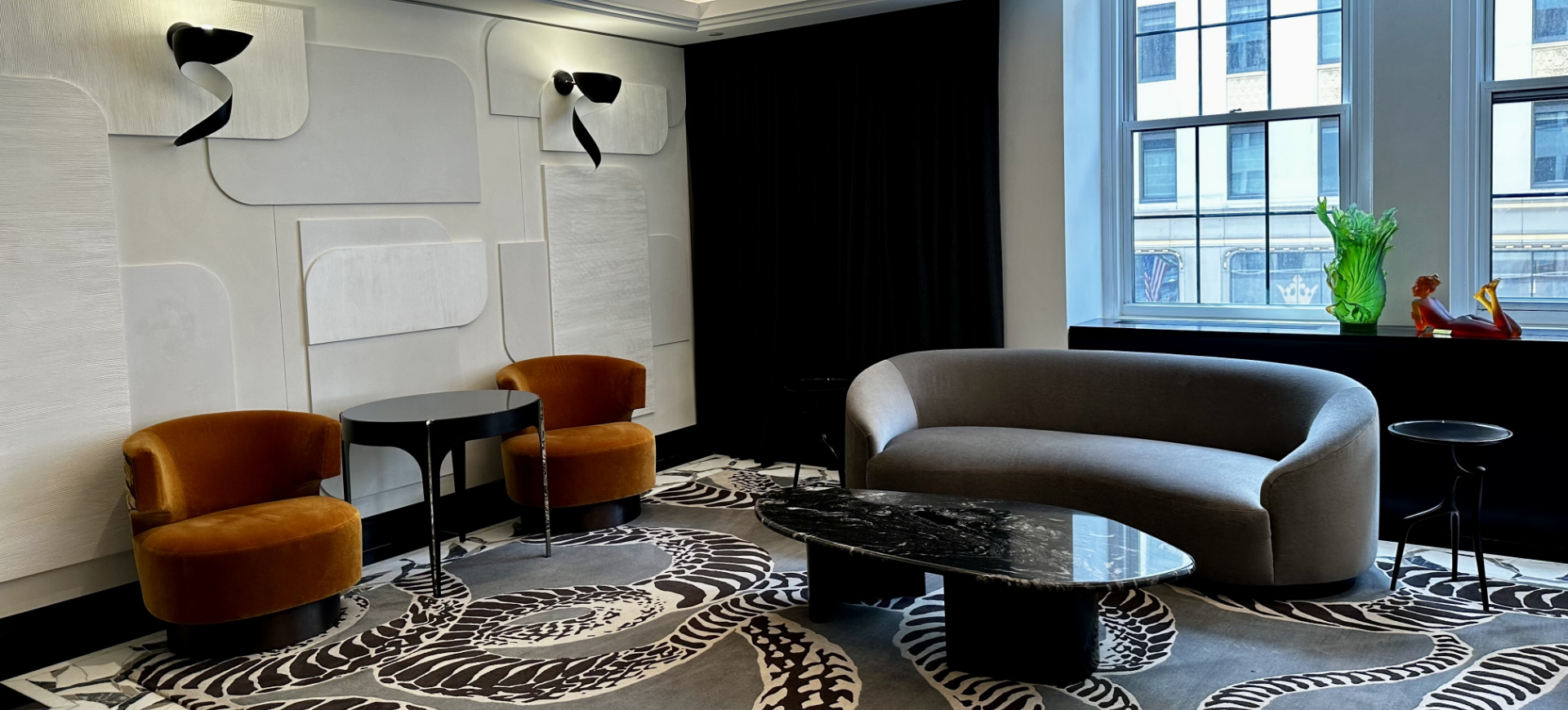 Modern living room with beige curved sofa, two orange velvet armchairs, black marble coffee table, black side table, large window with city view, black curtains, decorative sculptures and green glass vase on windowsill, and wall-mounted black and whi