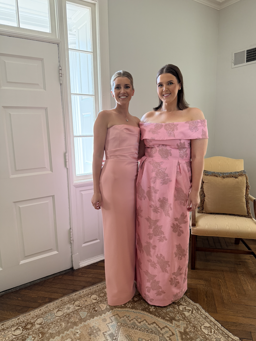 Two women in pink dresses standing indoors in front of a white door and window, smiling at the camera.