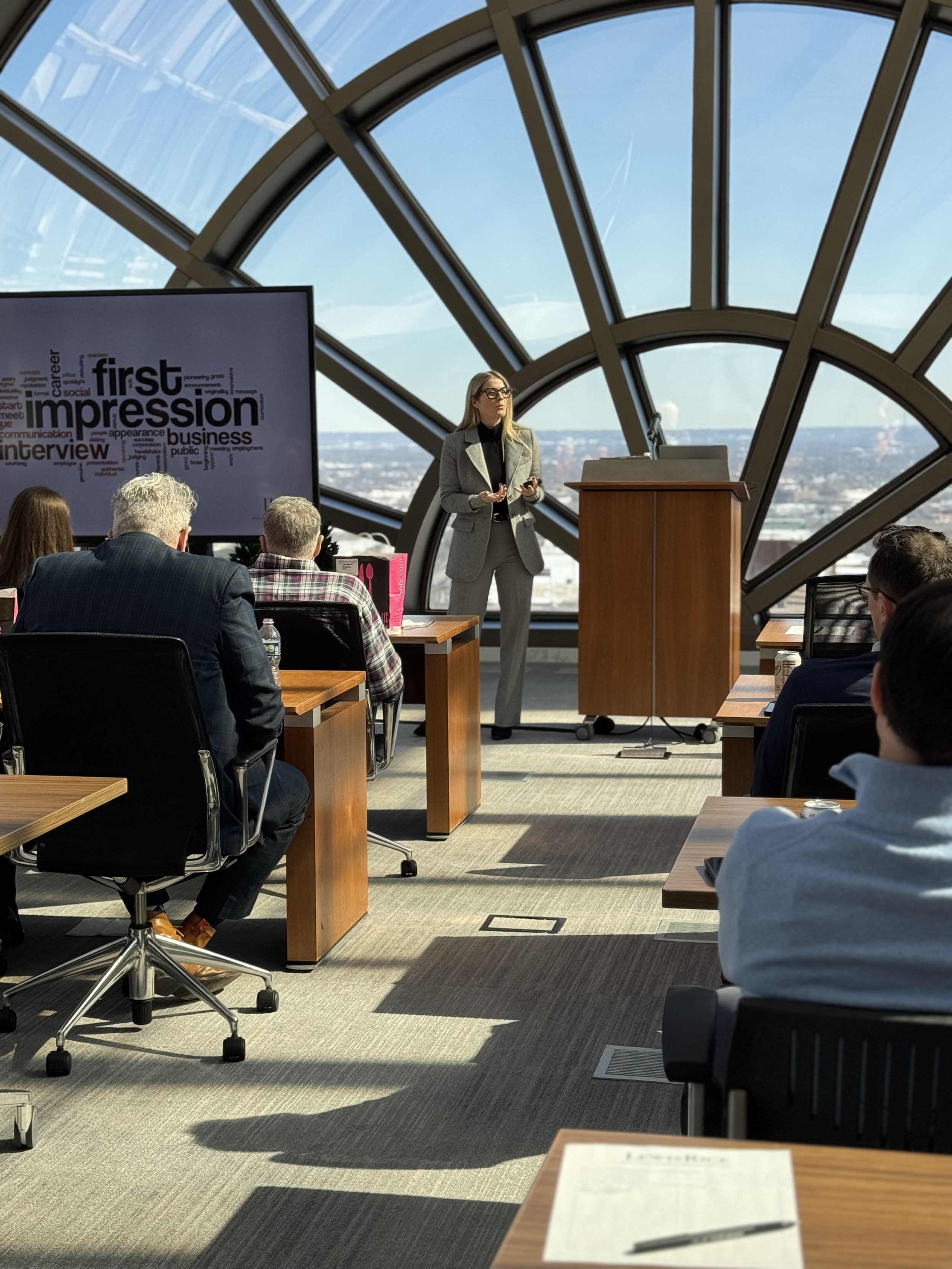 Laura K. Sawyier is giving a presentation to a group of people seated at tables in a conference room with large windows showing a cityscape and a blue sky.
