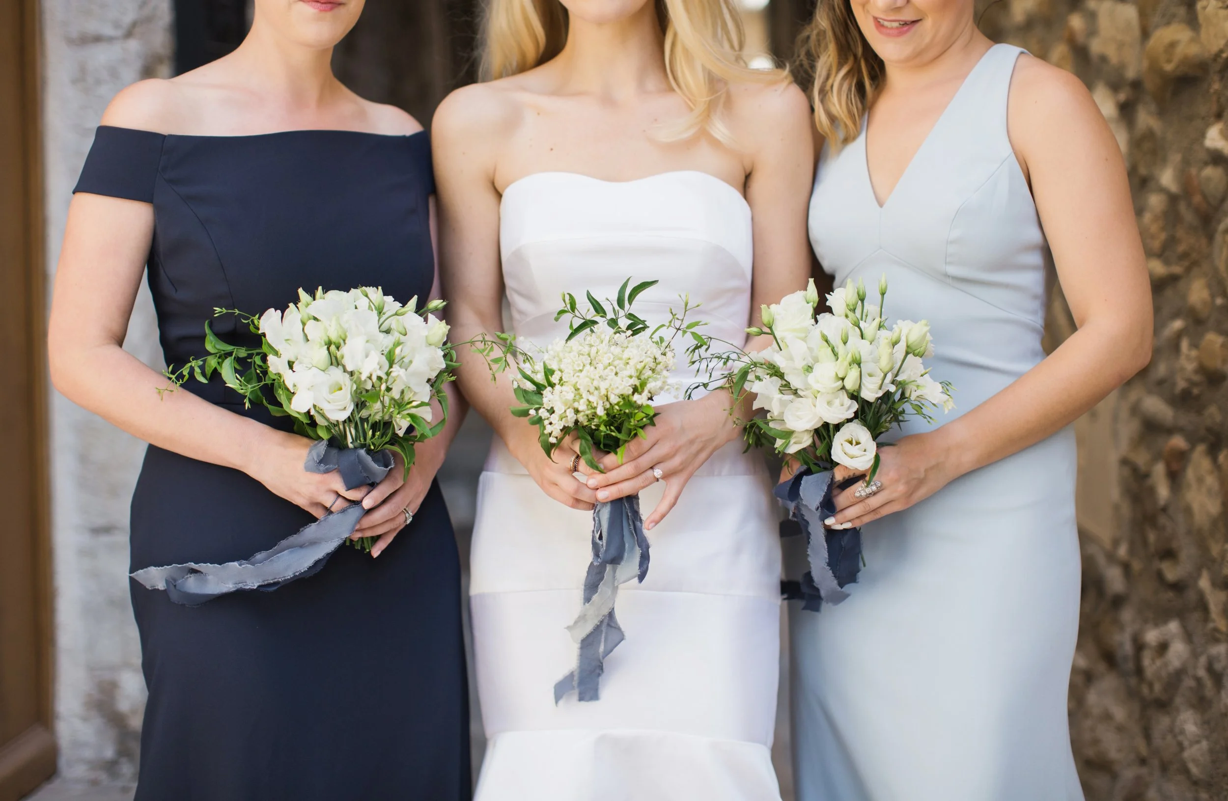 Three women in formal dresses holding bouquets of white flowers at a wedding.