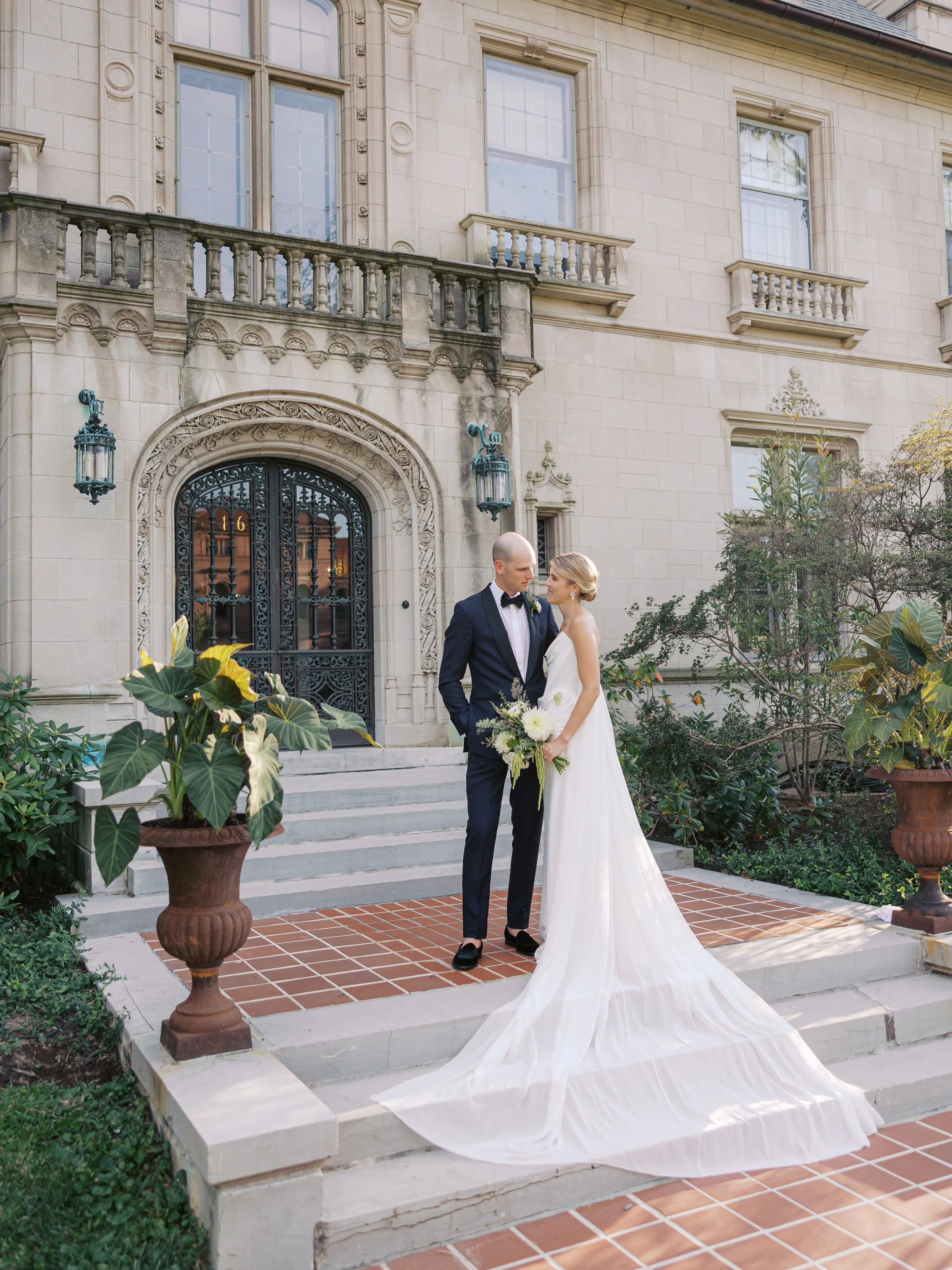 A bride and groom standing on the steps outside a historic building, looking into each other's eyes. The bride is in a long white dress with a train, holding a bouquet, and the groom is in a black tuxedo with a bow tie.