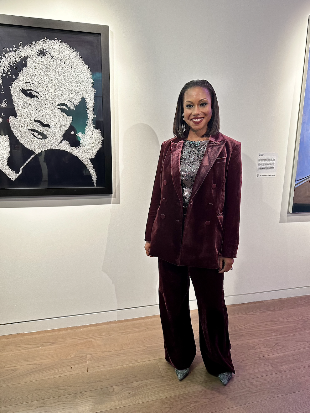 A woman in a burgundy velvet suit standing in an art gallery, smiling, with a portrait artwork made of small, shiny, reflective objects on the wall behind her.