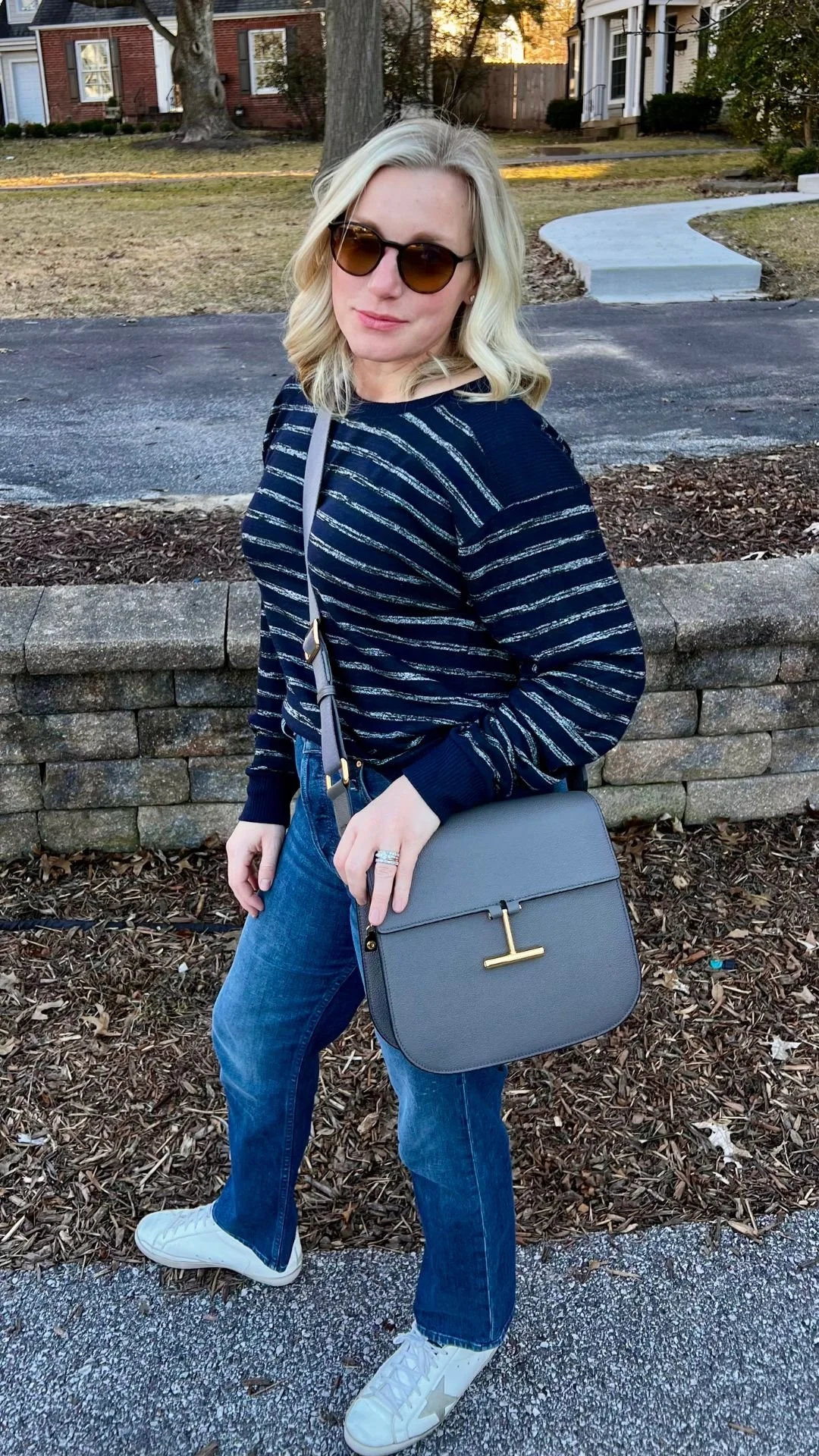 A woman with blonde wavy hair wearing sunglasses, a navy and silver striped sweater, blue jeans, and white sneakers, standing outdoors with a gray handbag, in front of a low stone wall and sidewalk in a residential neighborhood.