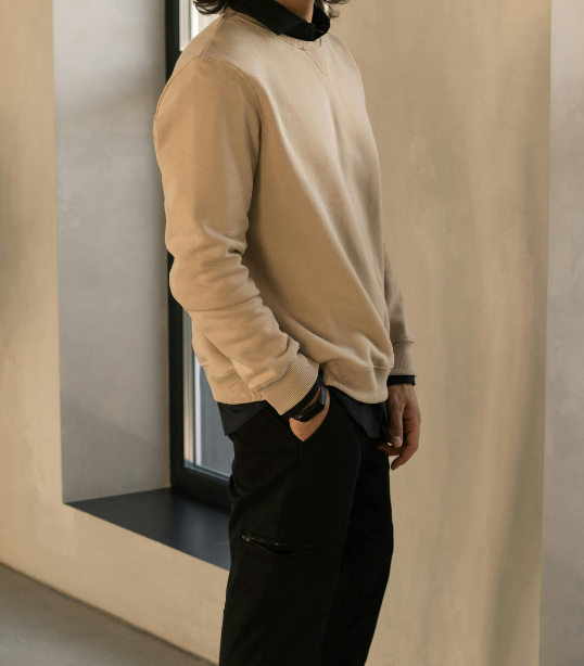 Person wearing a beige sweatshirt and black pants, standing indoors near a window.