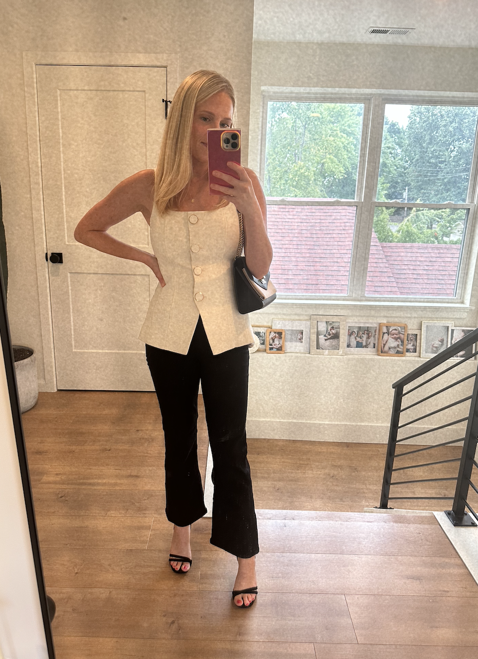 A woman taking a mirror selfie indoors, wearing a sleeveless cream-colored top with buttons and black pants, with a door and window in the background.