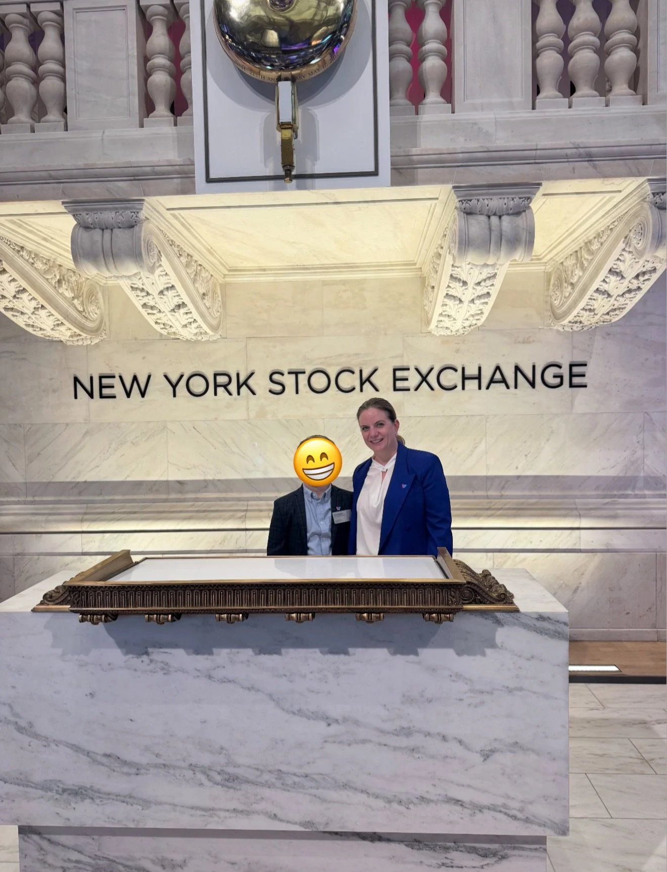Two people stand behind a marble counter at the New York Stock Exchange, with the exchange's name in large letters on the marble wall behind them. 