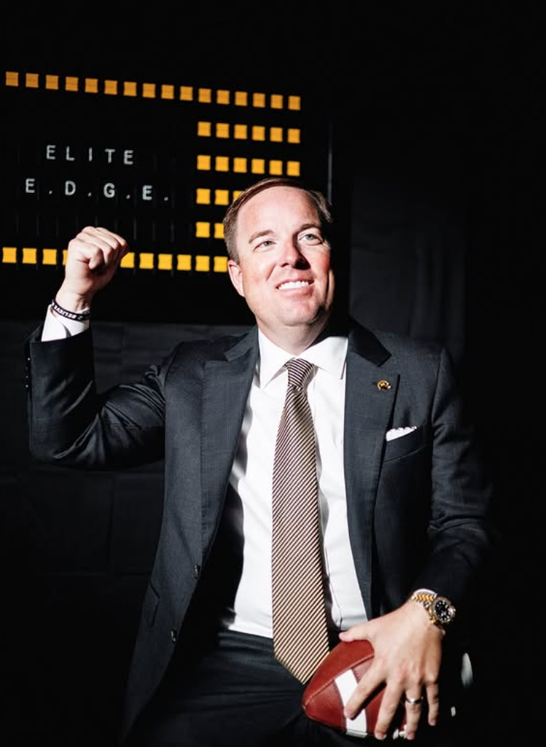 A man in a suit holding an American football, flexing his right arm in front of a scoreboard with yellow dots.