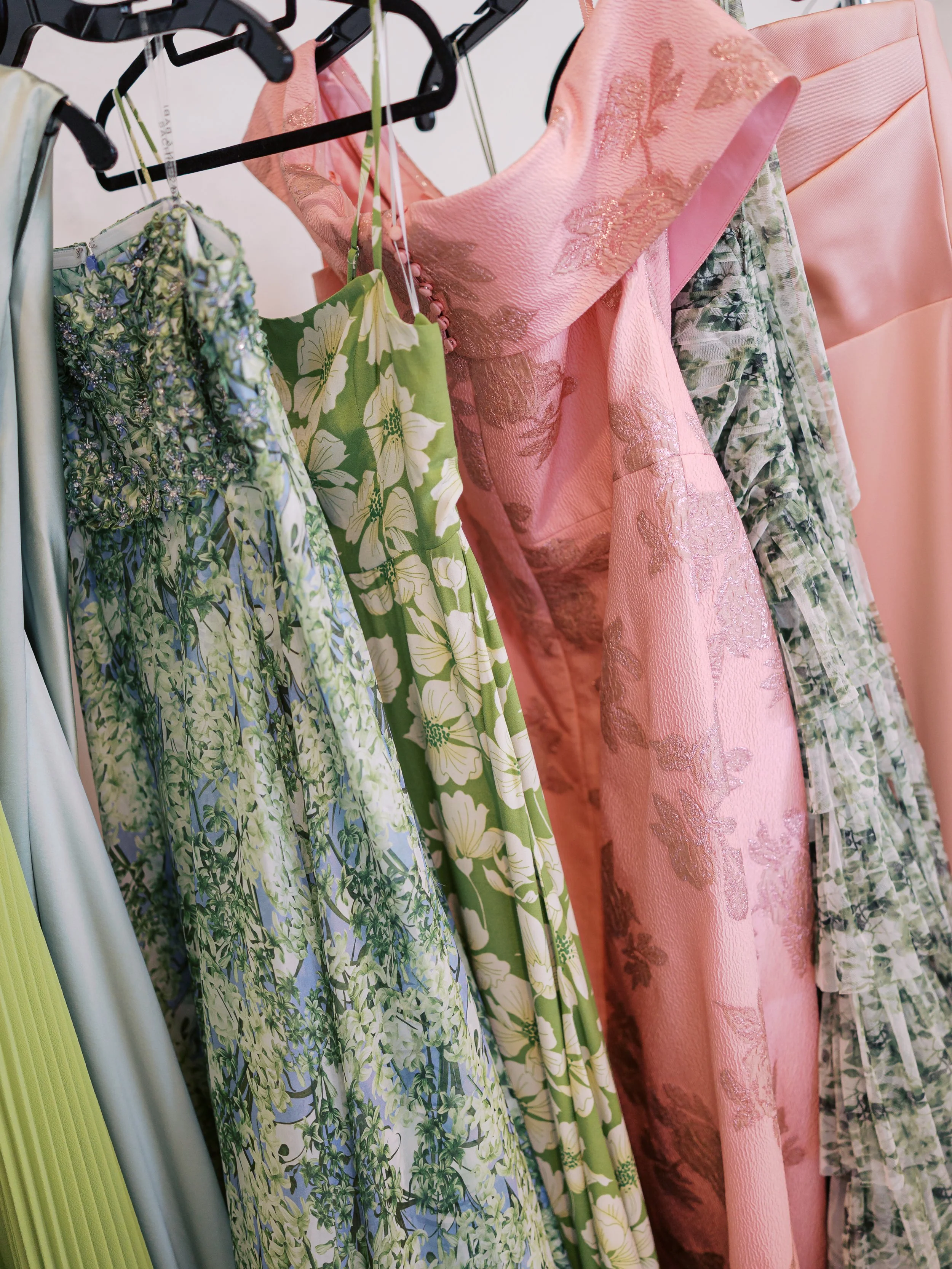 A variety of dresses hanging on a rack, featuring floral and pastel patterns with colors like green, pink, and cream.