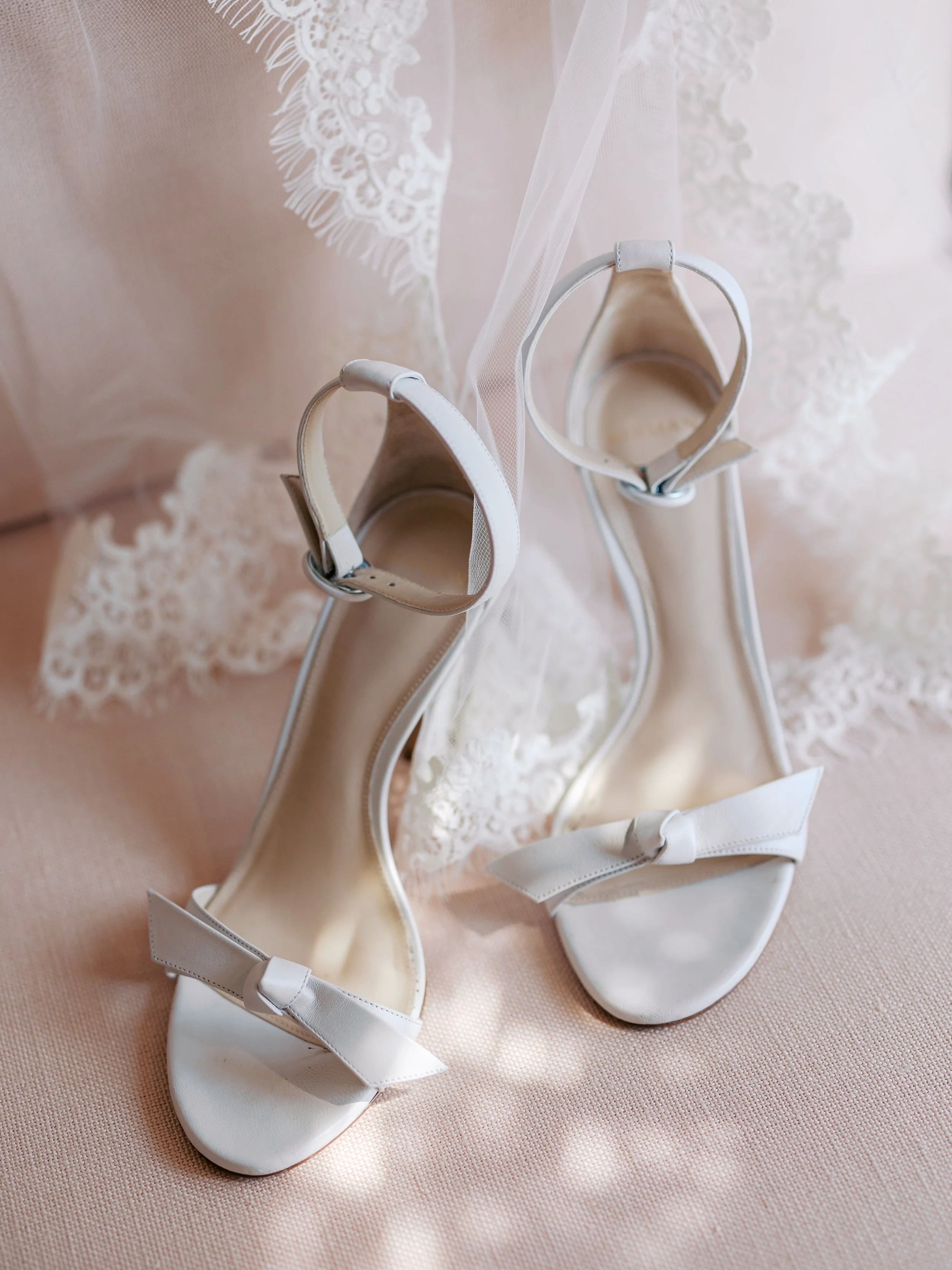 A pair of white high-heeled sandals with bows on the toe straps, placed on a pink fabric surface with lace fabric in the background.