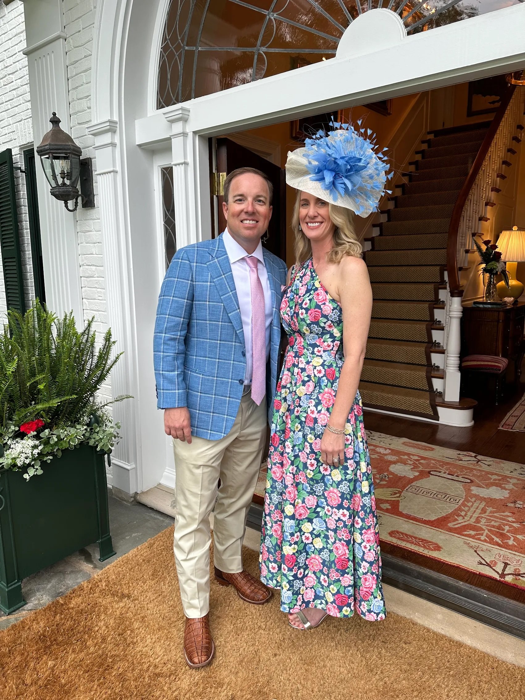 A man and woman standing inside the doorway of a house, smiling at the camera. The woman is wearing a colorful floral dress and a large blue hat, and the man is dressed in a blue checked blazer, pink tie, and beige pants.