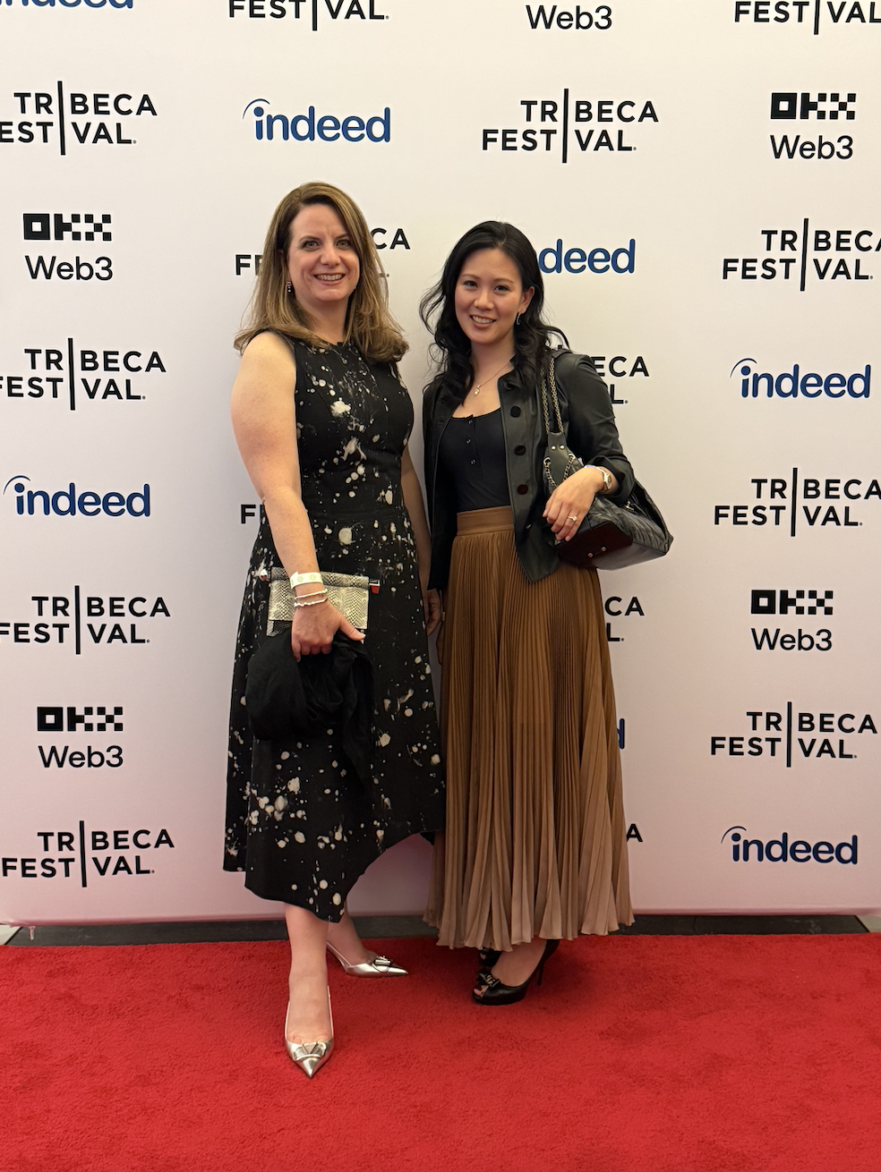 Two women standing on a red carpet at a Tribeca Festival event, smiling at the camera. One is wearing a black speckled dress and silver heels, and the other is wearing a black jacket and a pleated brown skirt, carrying a black purse.