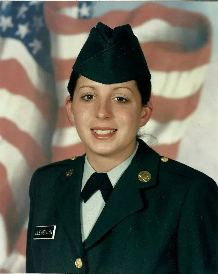 A young woman in a formal military uniform with a name tag that reads 'LLEWELLYN' poses for a portrait in front of an American flag backdrop.