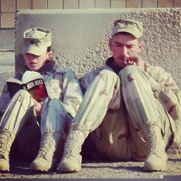 Two soldiers in uniform in the desert with backpacks and caps sitting on the ground against a wall, one reading a book and the other smoking.