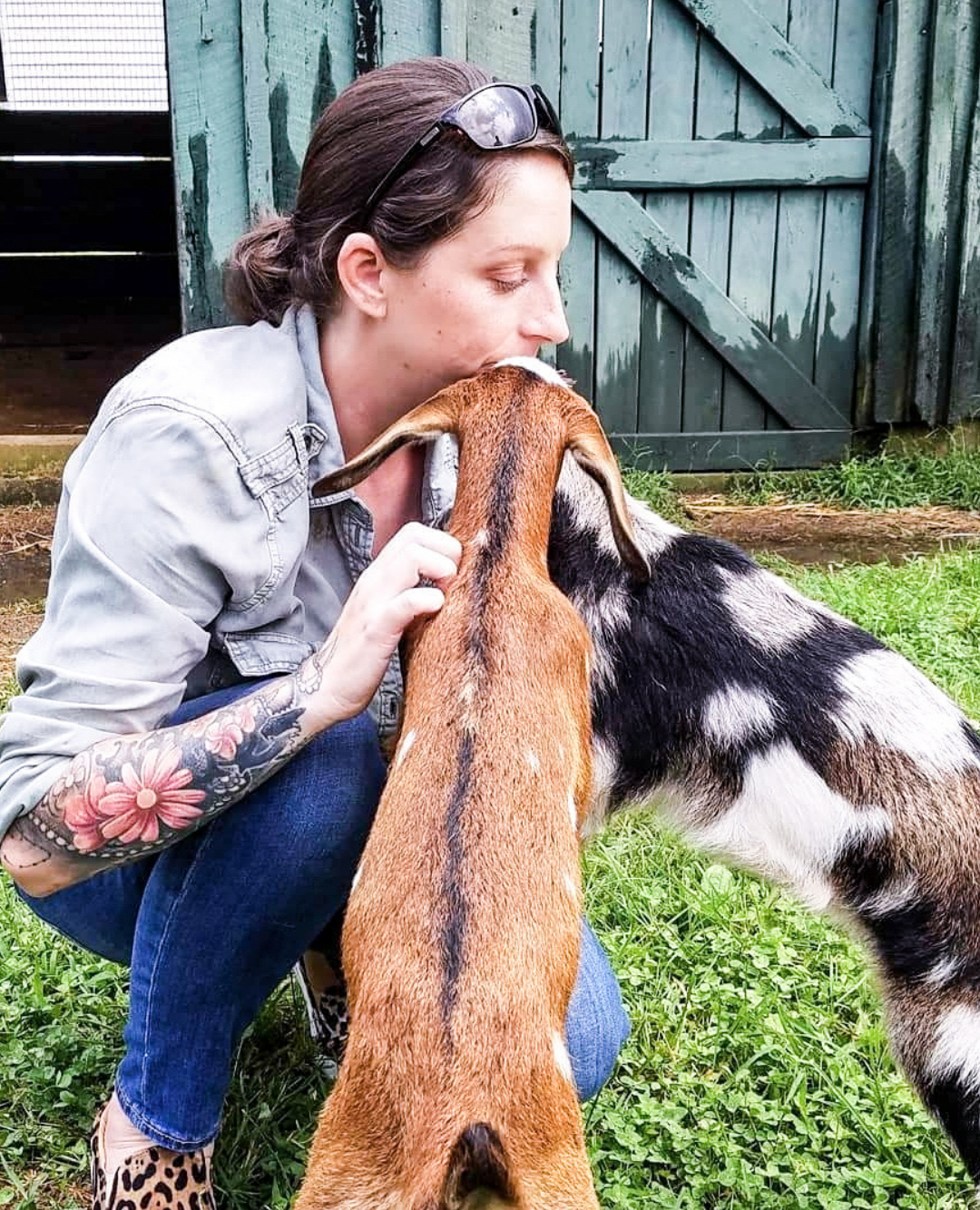 A woman with tattoos and sunglasses resting on her head kneeling in front of a goat and a dog outside near a green wooden shed, with the woman gently holding the goat's head.