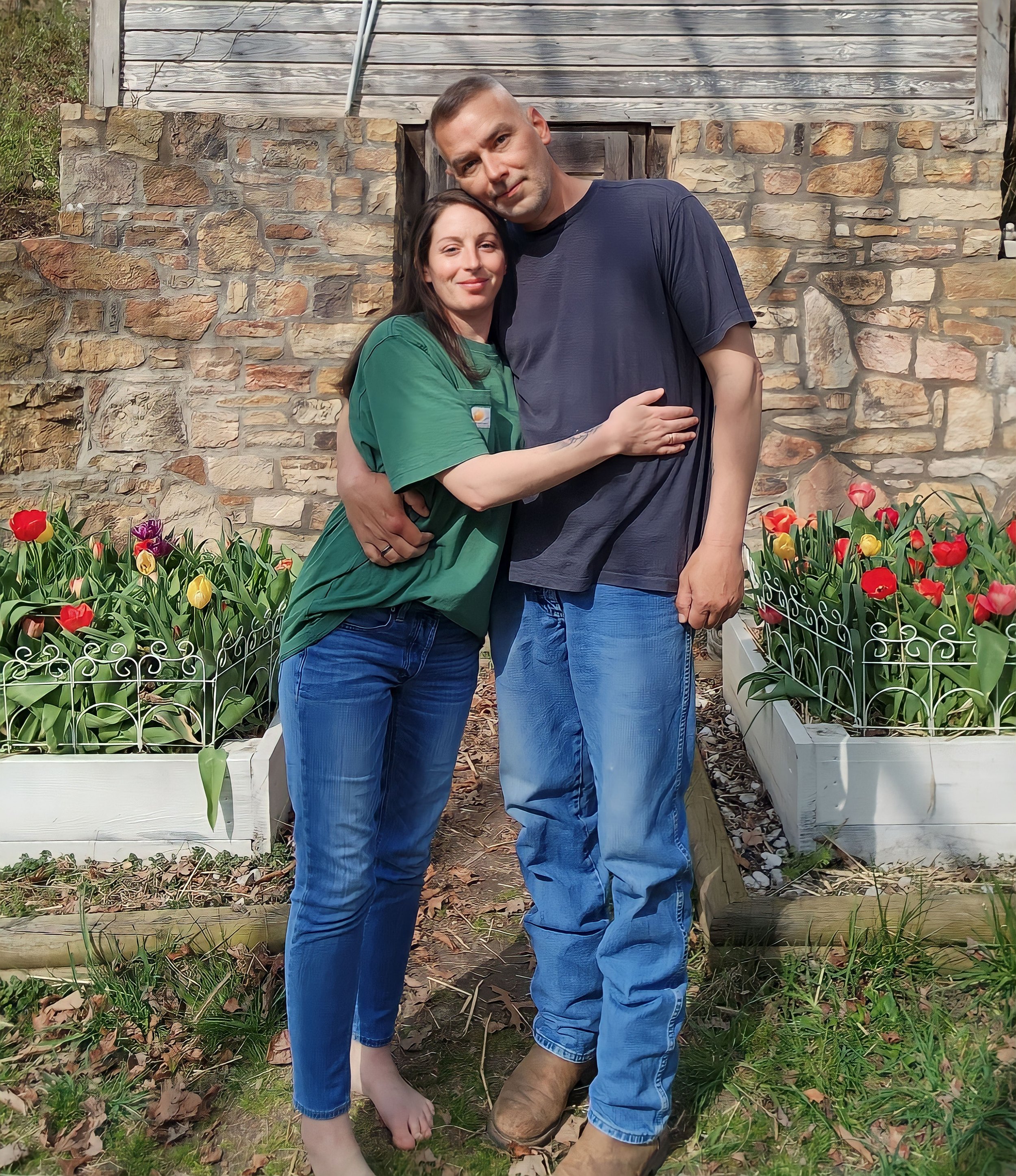 A man and woman stand close together outdoors, hugging, with tulips in a garden bed behind them and a rustic stone and wood building in the background.