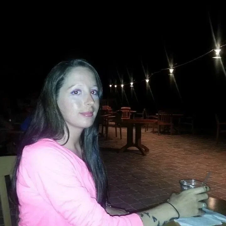 A woman with long dark hair and purple eye makeup sitting at a table outdoors at night, holding a glass container with a spoon.