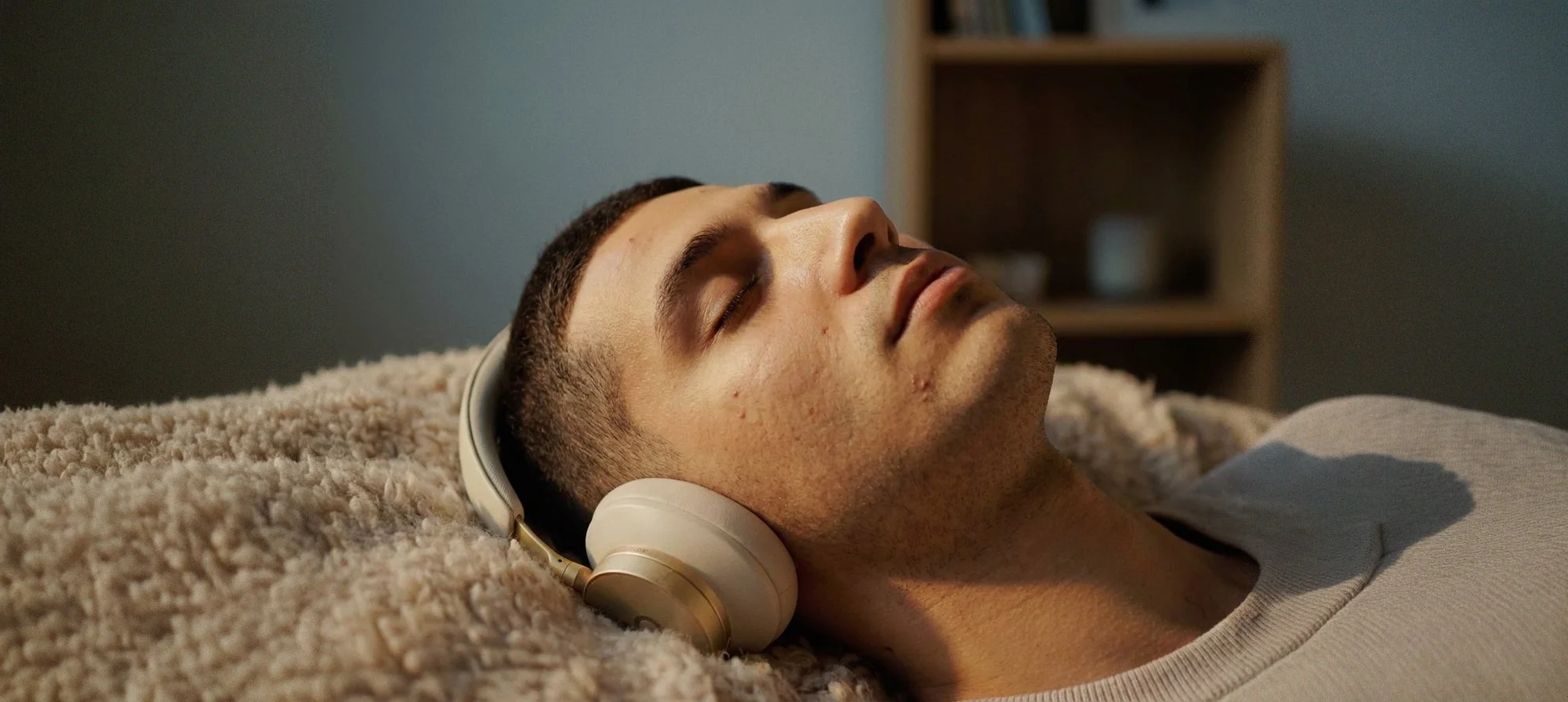 A young man with short dark hair and facial acne is lying on a bed with a beige plush blanket, wearing white headphones, with his eyes closed in a relaxed state.