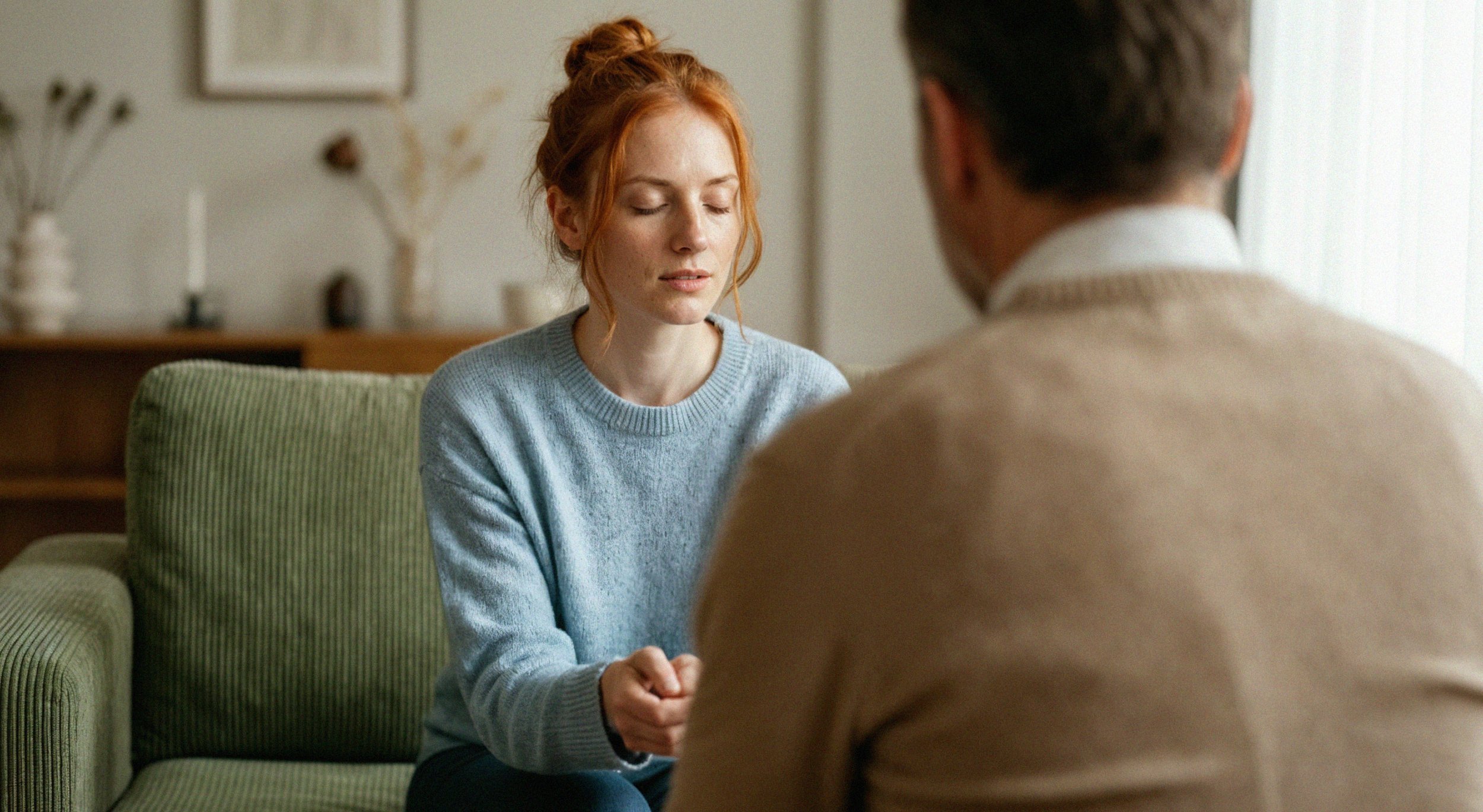 A woman with red hair in a bun sitting on a green couch with her eyes closed, holding hands with a man who has gray hair and is wearing a beige sweater, in a cozy living room.