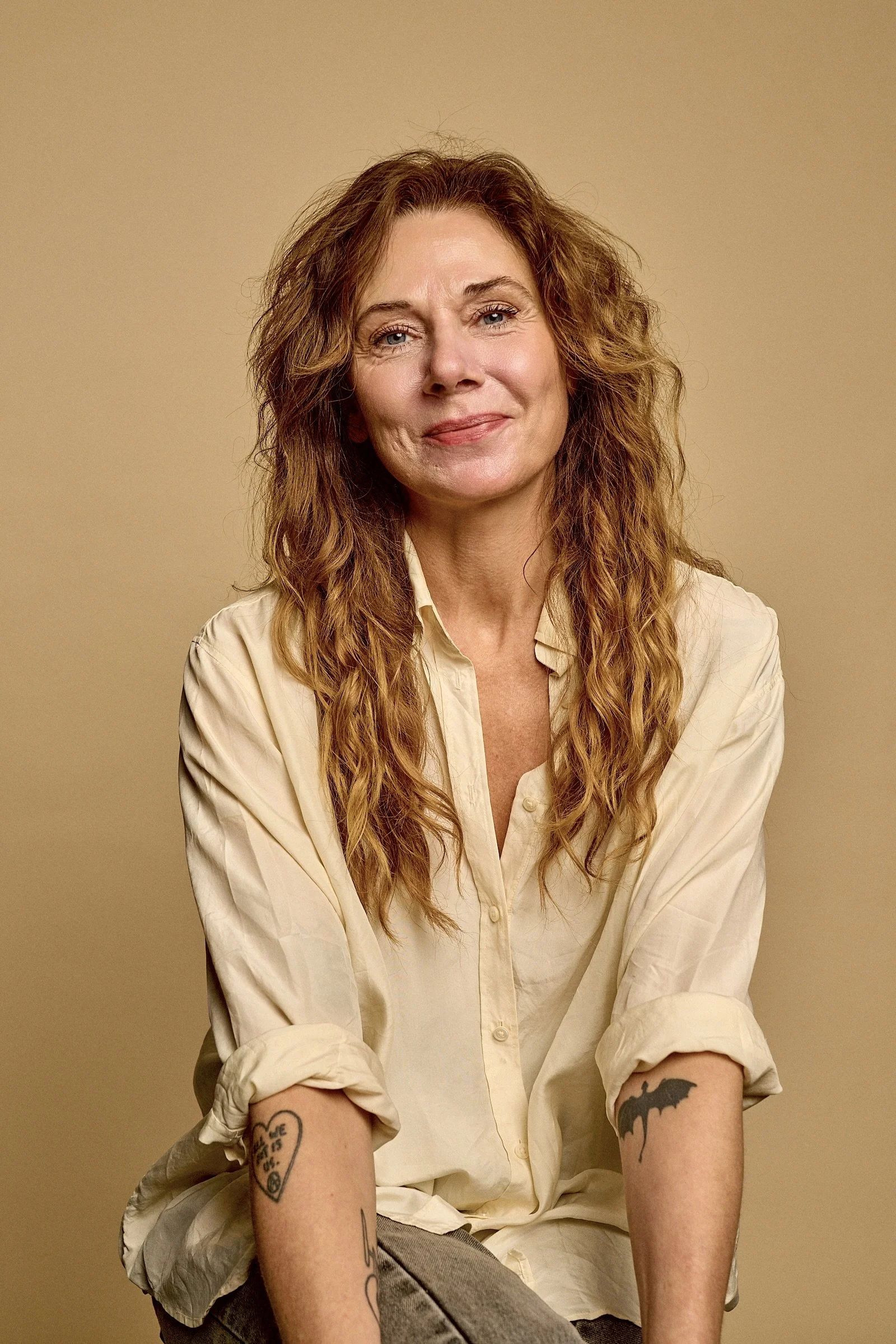 A woman with long wavy red hair and tattoos on her arms, wearing a beige shirt with rolled-up sleeves, sitting against a plain yellow wall, smiling gently at the camera.
