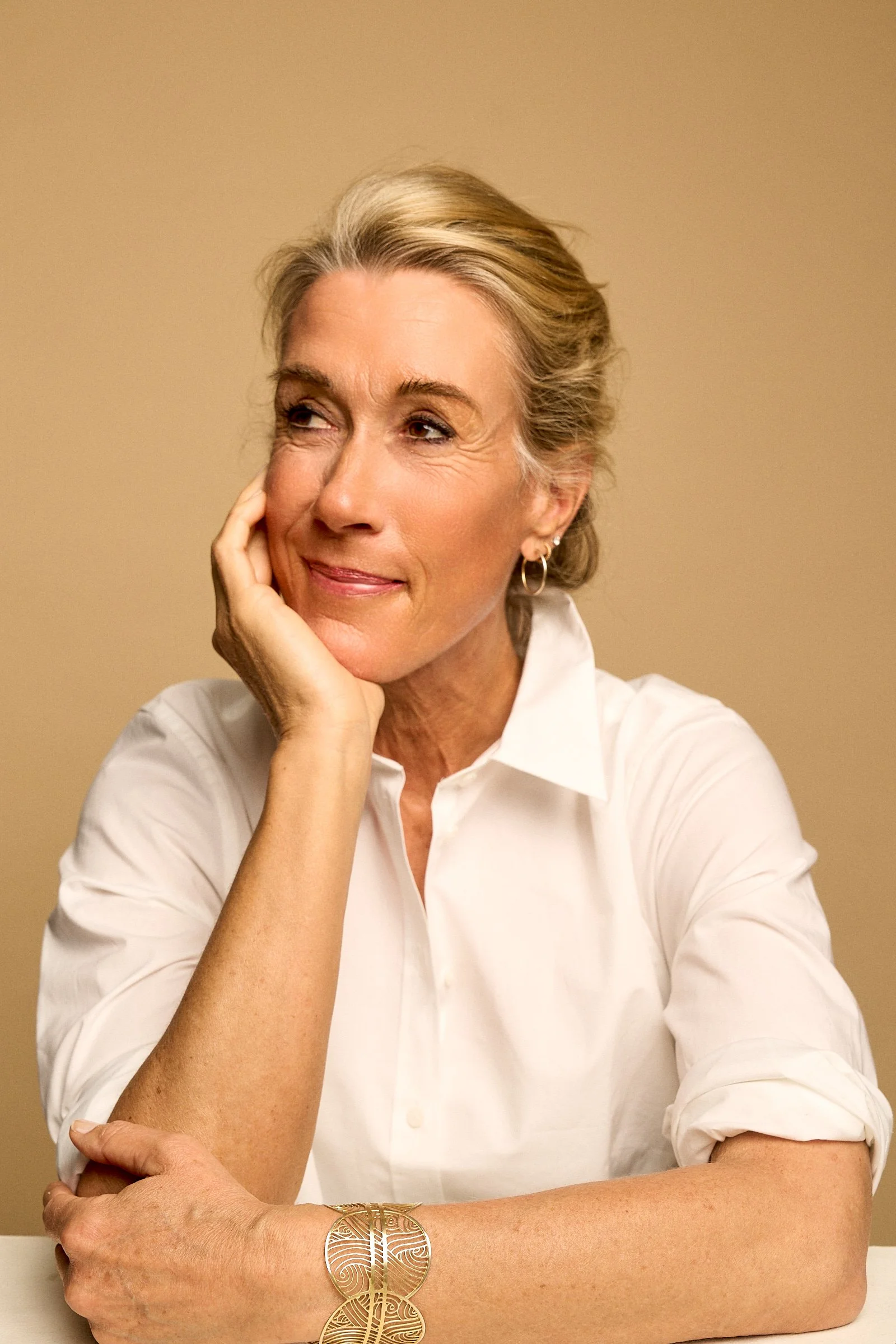 A mature woman with blonde hair styled back, wearing a white shirt and gold jewelry, resting her chin on her hand and looking to her right against a beige background.