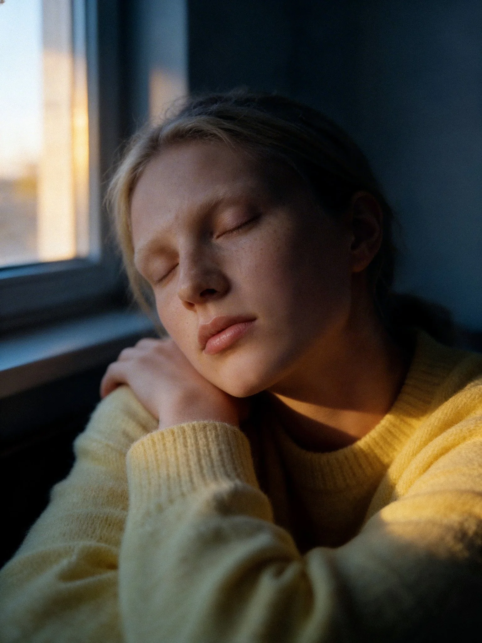 A young person with light skin and blonde hair peacefully sleeping by a window, wearing a yellow sweater.