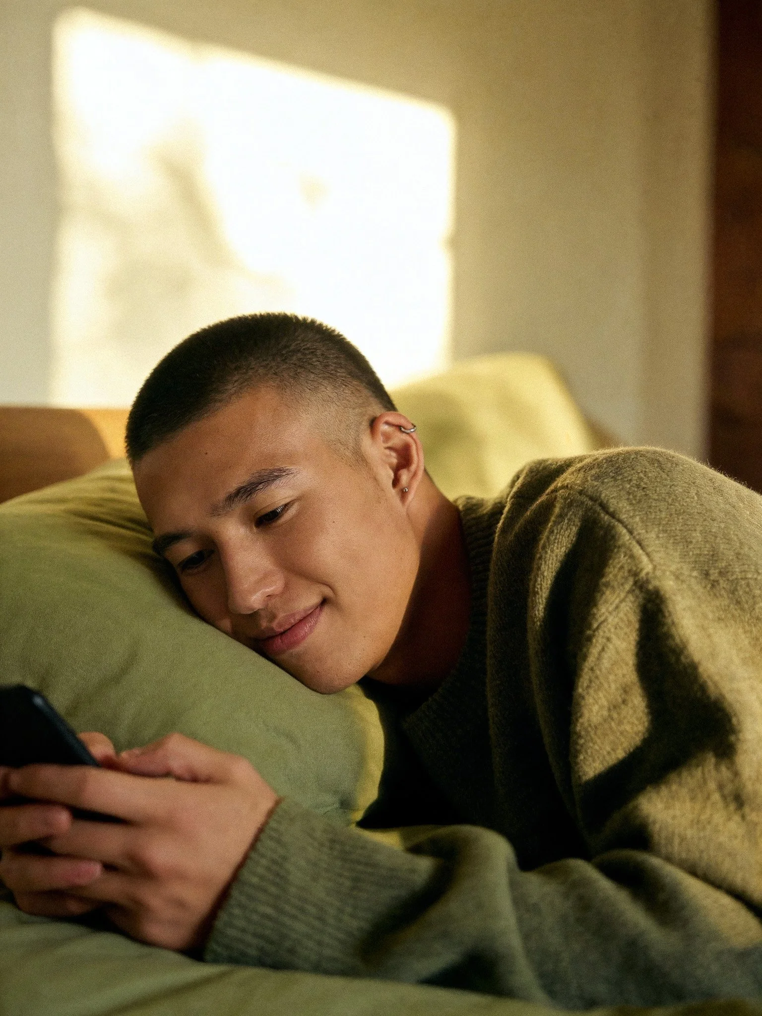 Young man with short hair and earrings, lying on a yellow pillow, looking at his phone in a cozy room with sunlight streaming through a window.