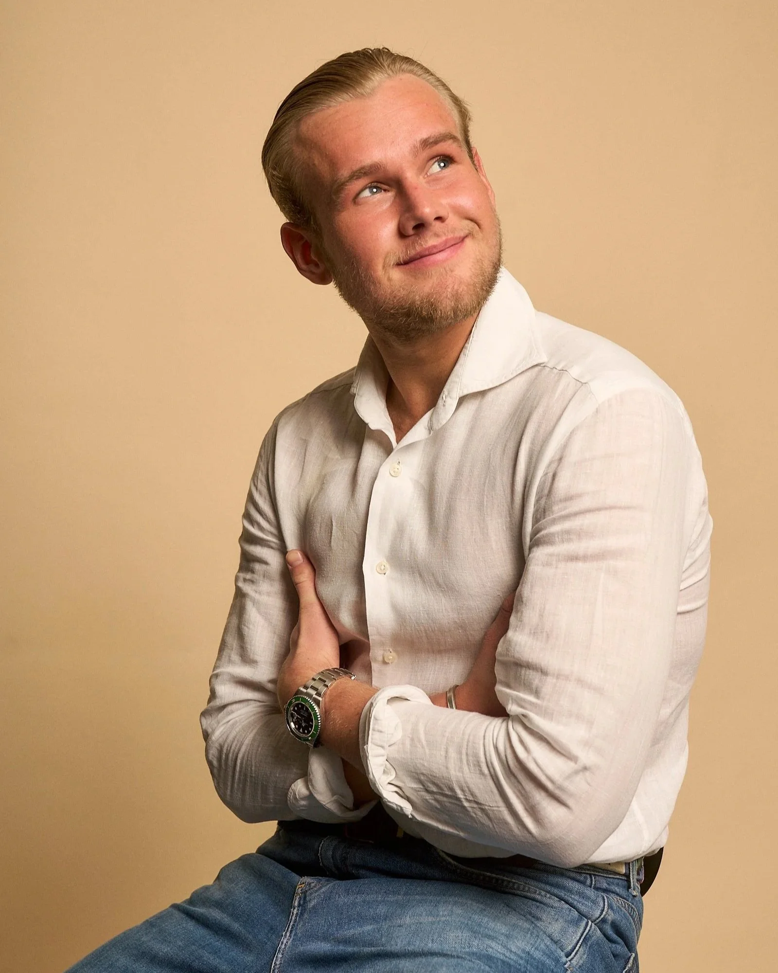 A man with blonde hair and a beard, smiling while looking up and to the right. He is wearing a white shirt and a silver watch, with a woman’s hand gently touching his arm. The background is plain beige.