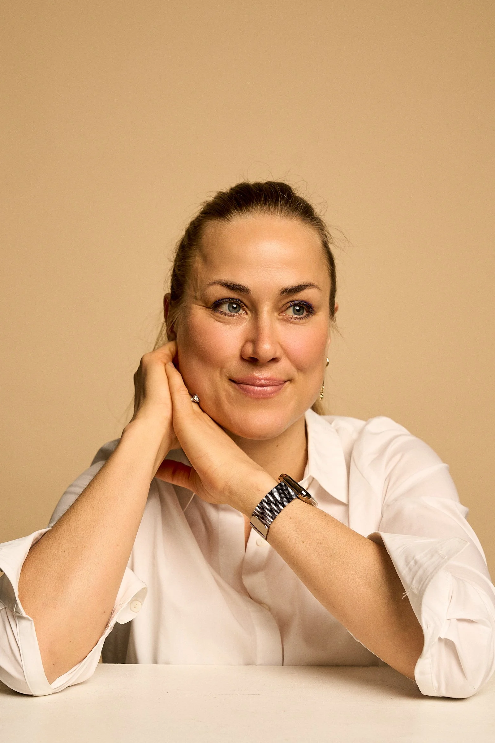 A woman with blonde hair styled in a bun, wearing a white shirt, sitting at a table with her hands resting on her face, smiling softly, against a beige background.