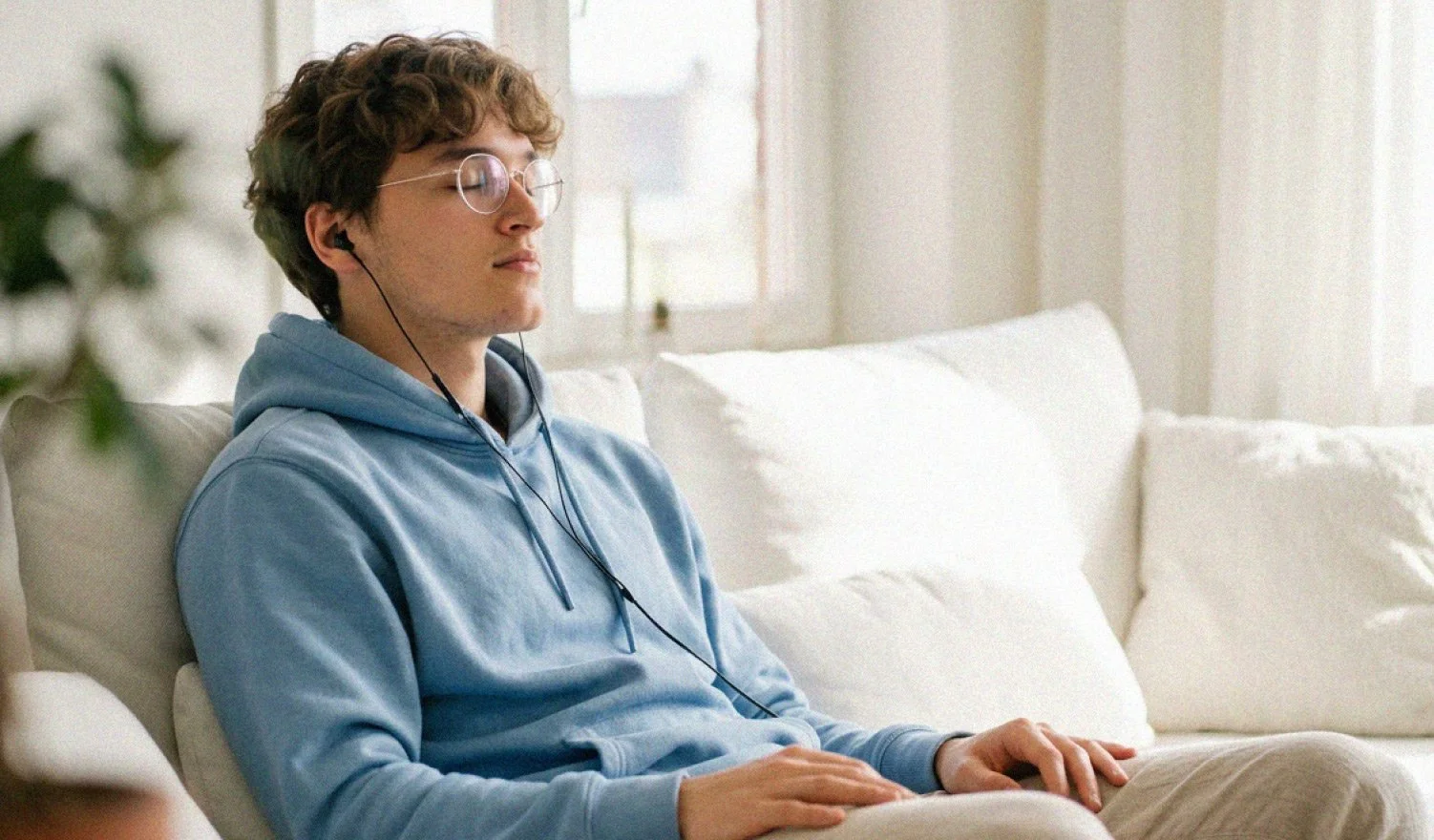 Young man with curly hair and glasses, wearing a blue hoodie, sitting on a white couch with closed eyes and earphones.