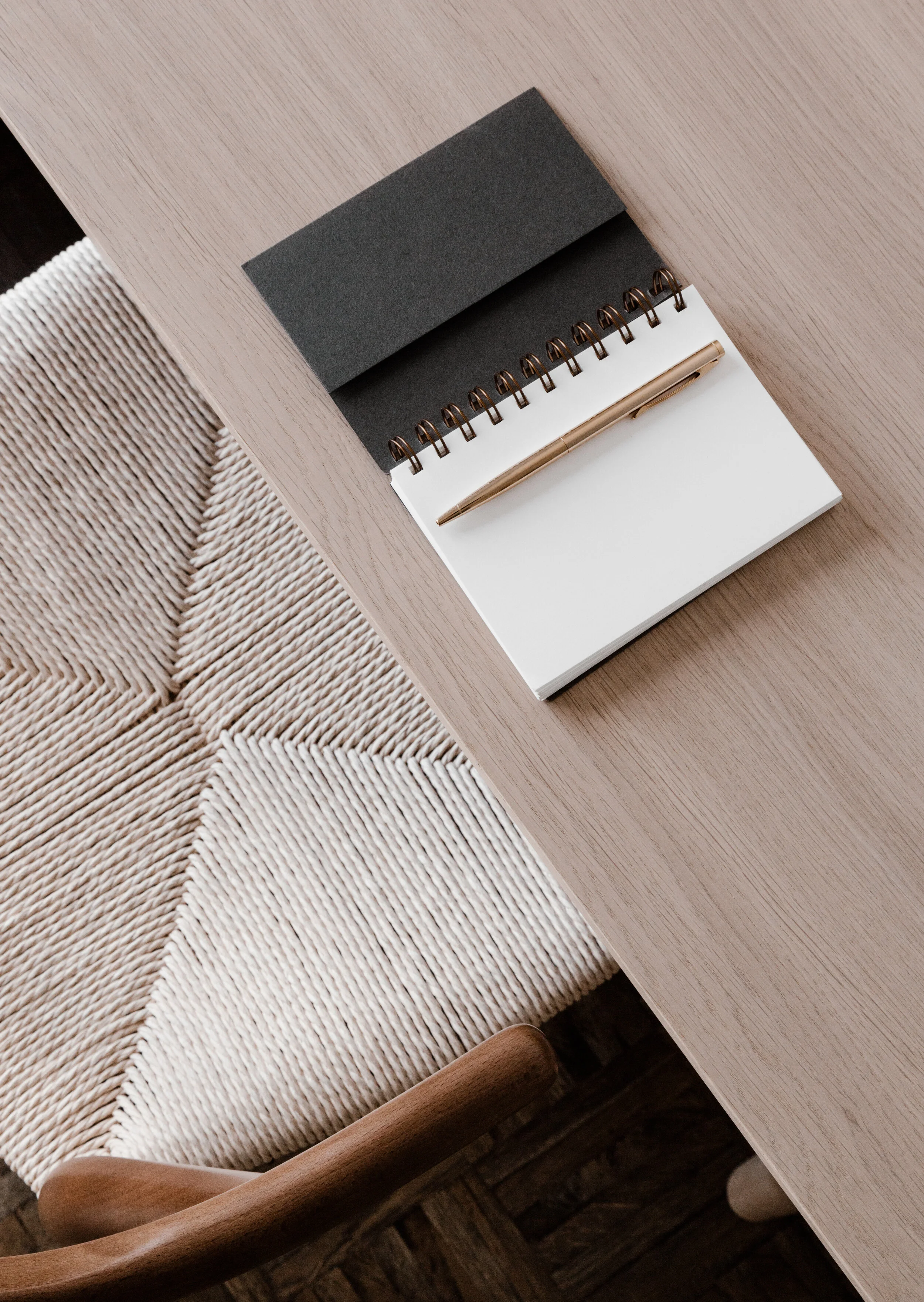 A spiral notepad with a black cover, a white notepad, and a gold pen on a light wooden desk next to a woven chair.
