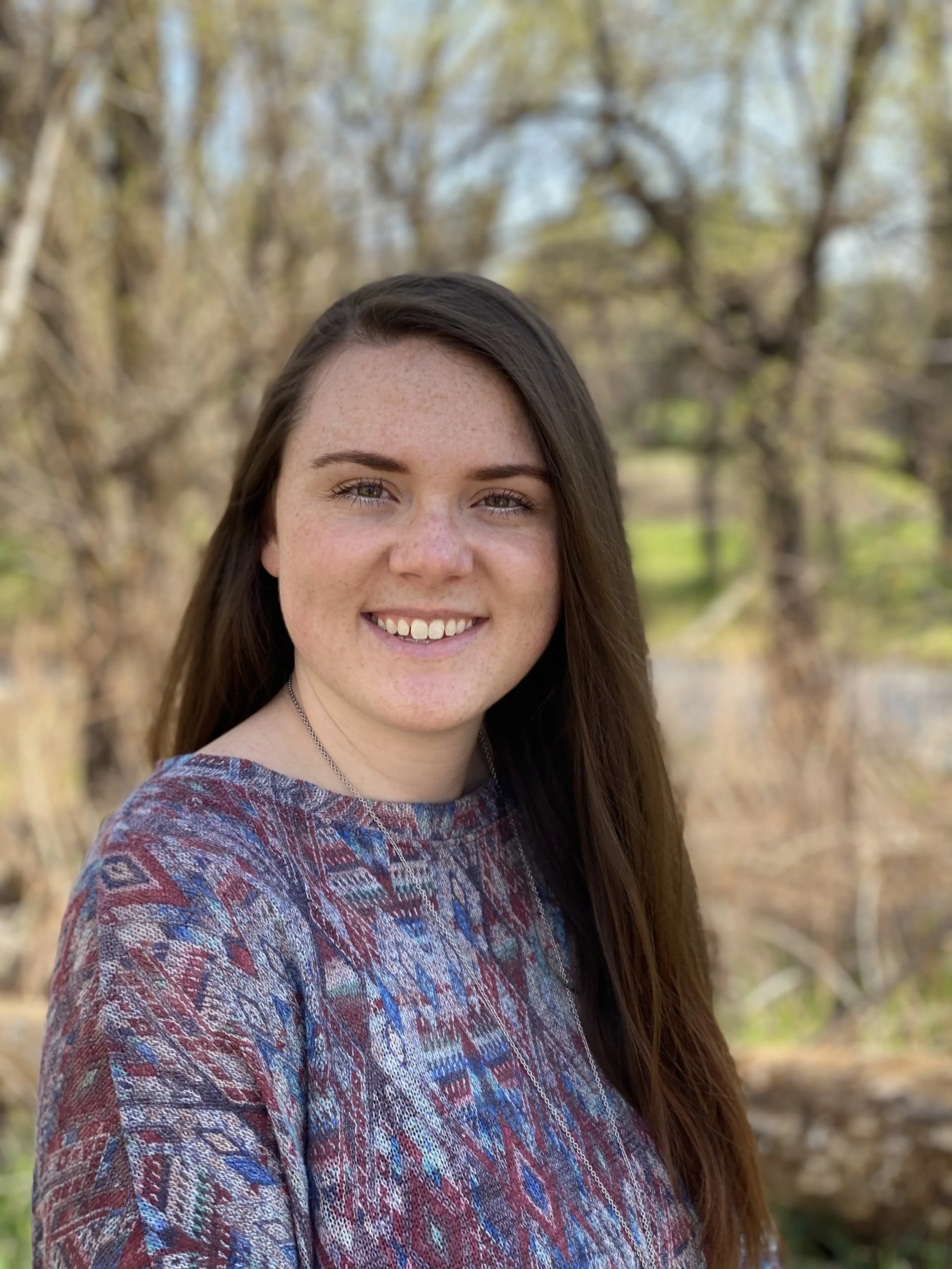 A smiling woman with long brown hair and light skin standing outdoors in front of trees with budding leaves during spring.