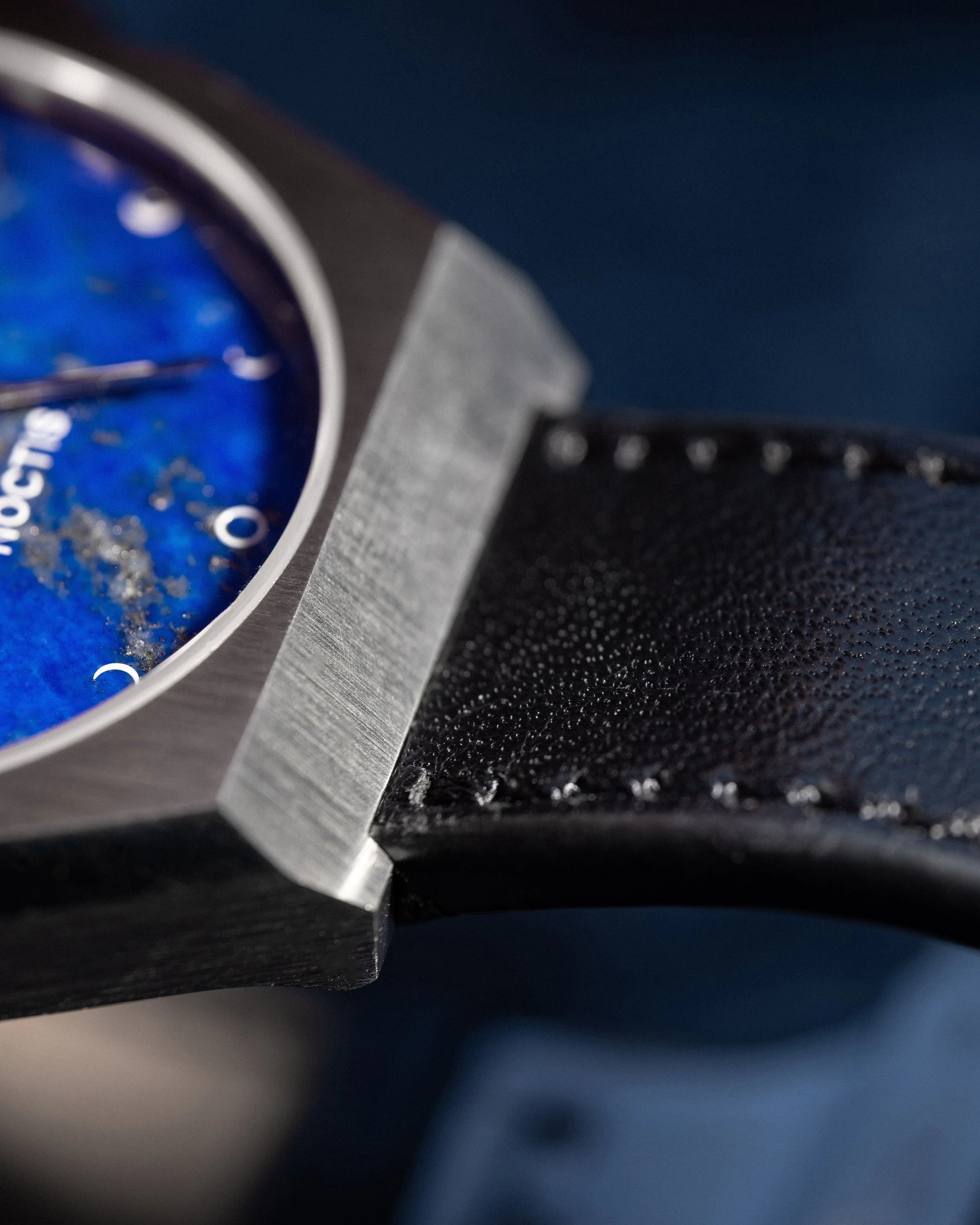 Nahaufnahme einer Uhr mit schwarzem Lederarmband und silbernem Gehäuse, teilweise sichtbar, auf dunklem Hintergrund.