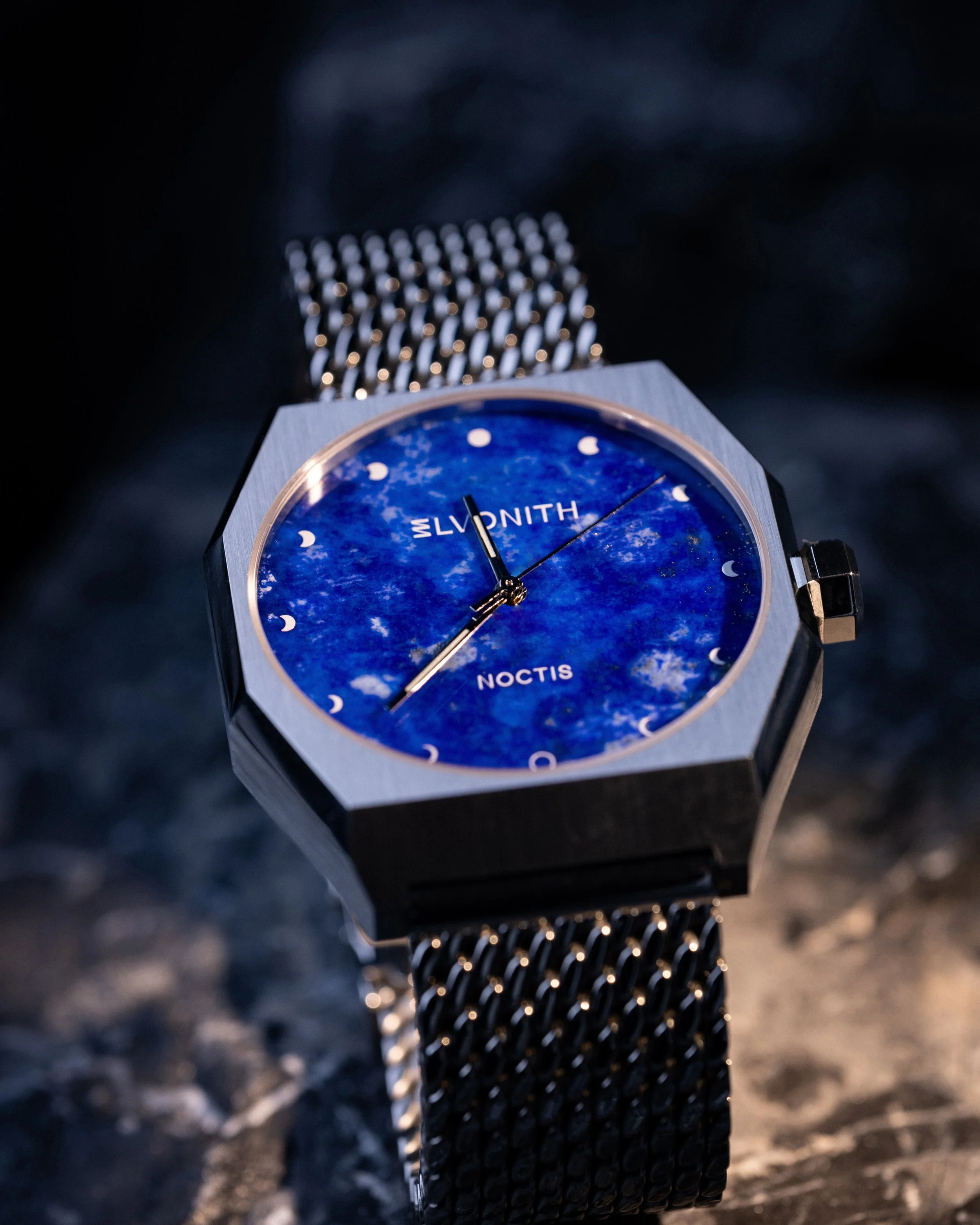 Eine Armbanduhr mit blauem Zifferblatt, Indizes als Mondphasen, und Edelstahl-Gehäuse auf dunklem Hintergrund.