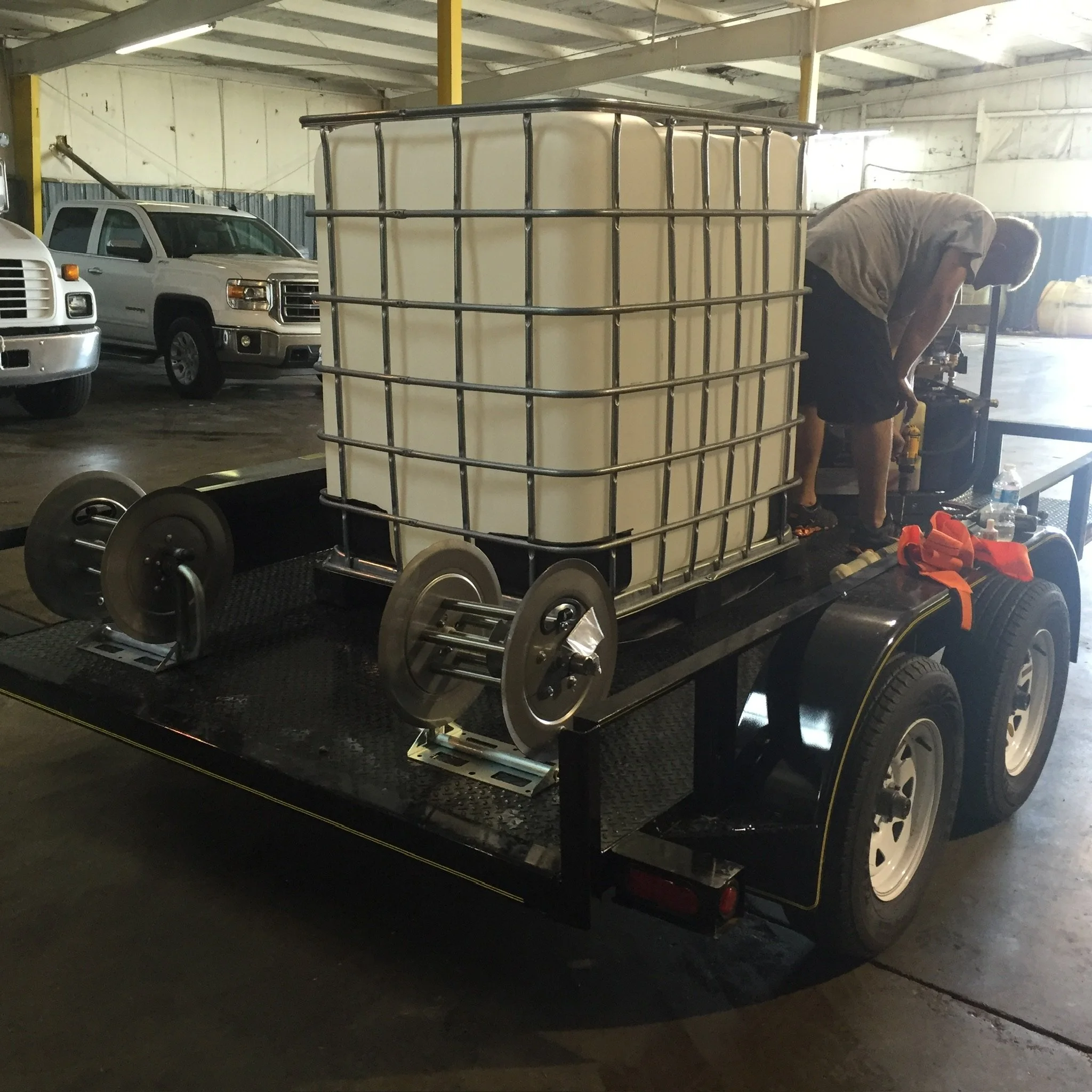 Person working on a utility trailer with a large storage tank and hoses indoors.